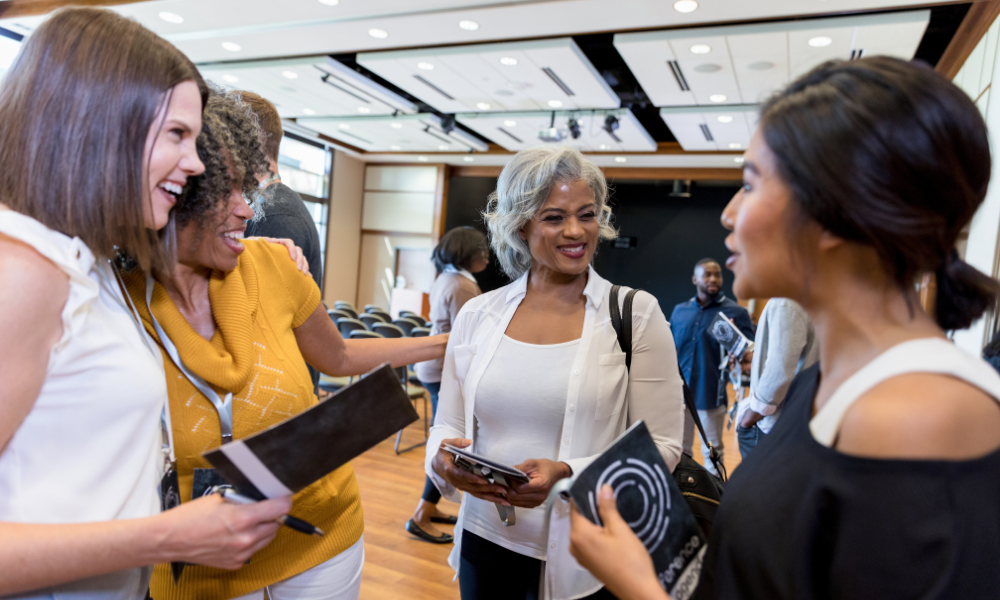 Four women engaged in conversation at a conference or seminar, smiling and holding brochures or programs, in a well-lit room with rows of chairs.