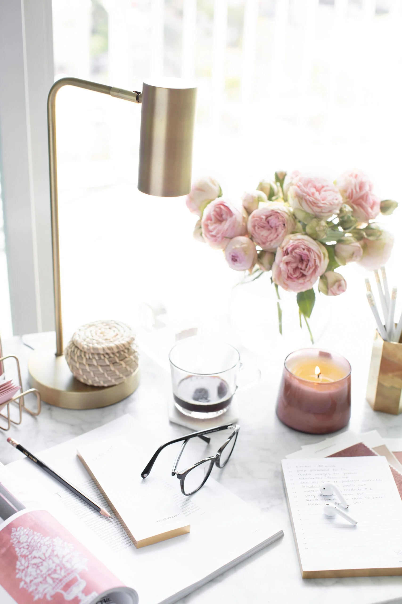 A white marble desk with pink and white flowers in a vase, a candle, a glass of dark beverage, notebooks, glasses, a pencil, and a pair of wireless earbuds.
