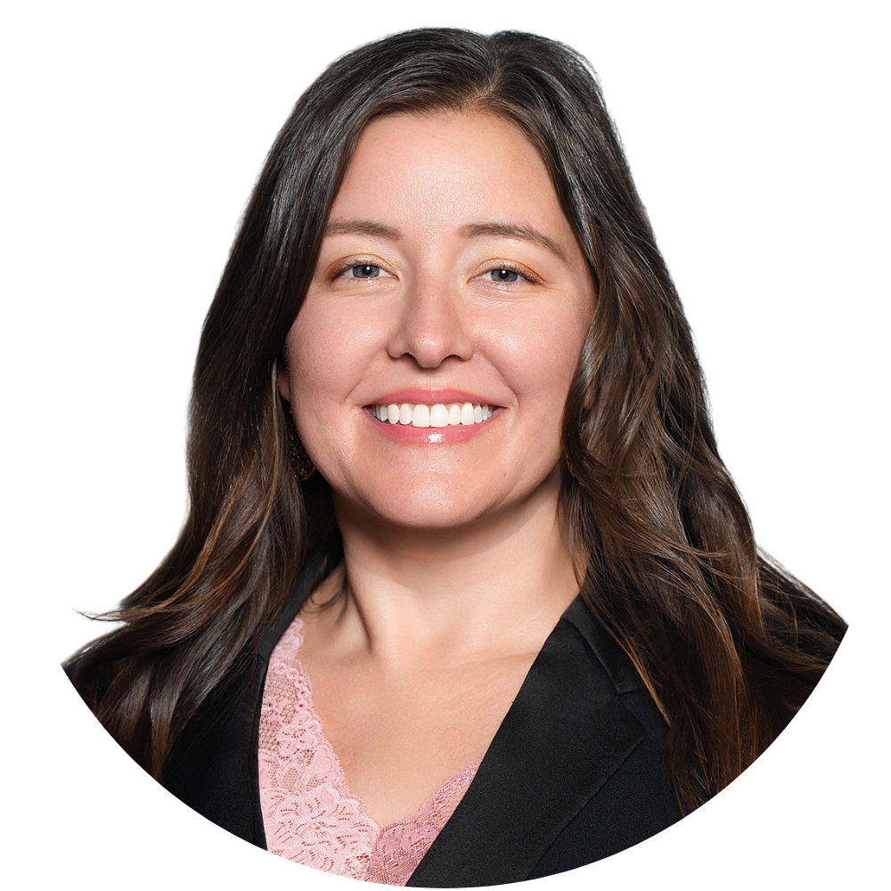 A woman with long brown hair, smiling against a white background, wearing a black blazer over a pink lace top.