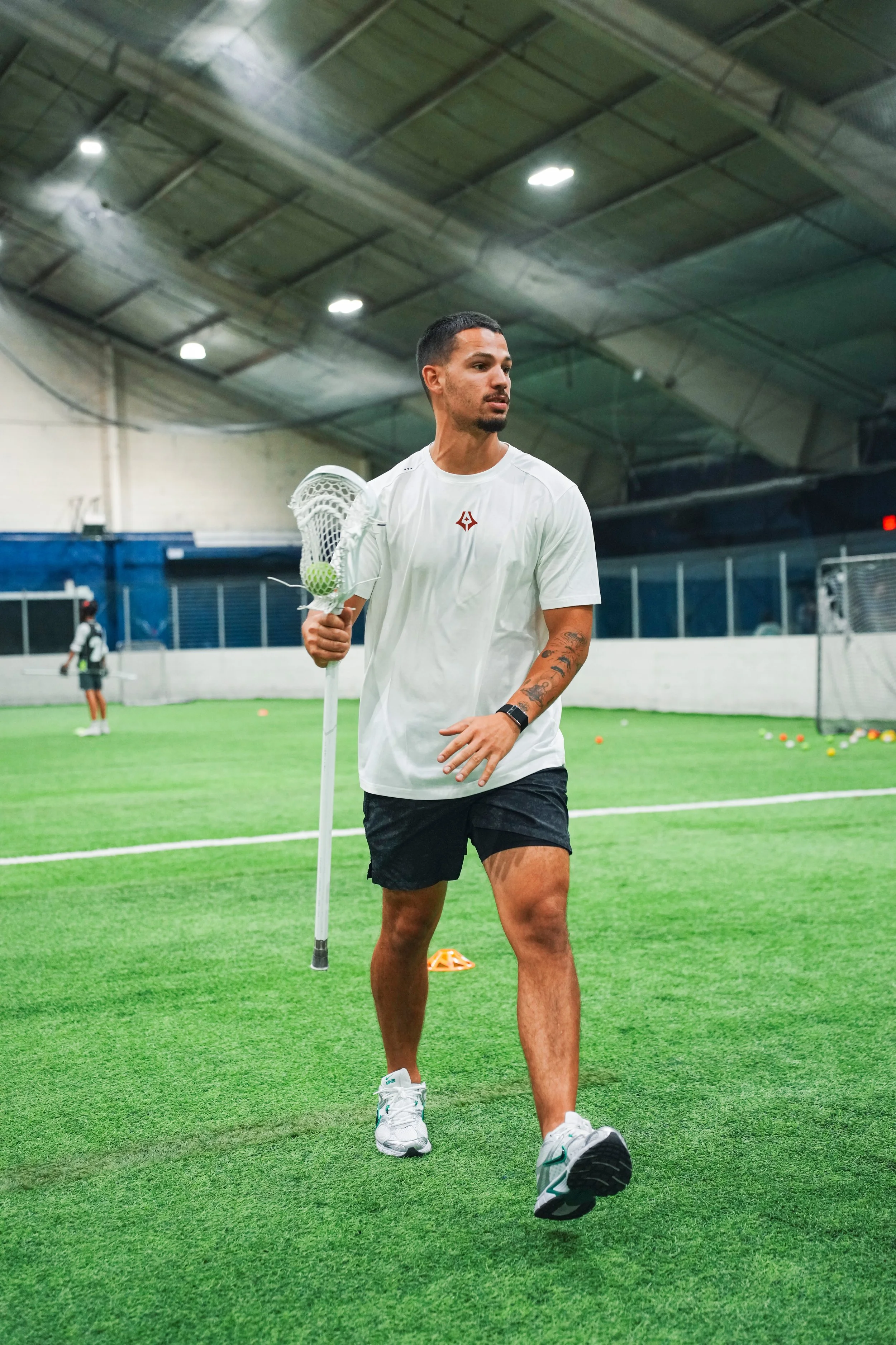 A man walking on an indoor turf field holding a lacrosse stick with a ball in the net. In the background, there is another person also with a lacrosse stick. The ceiling is high with visible lights and structural beams.