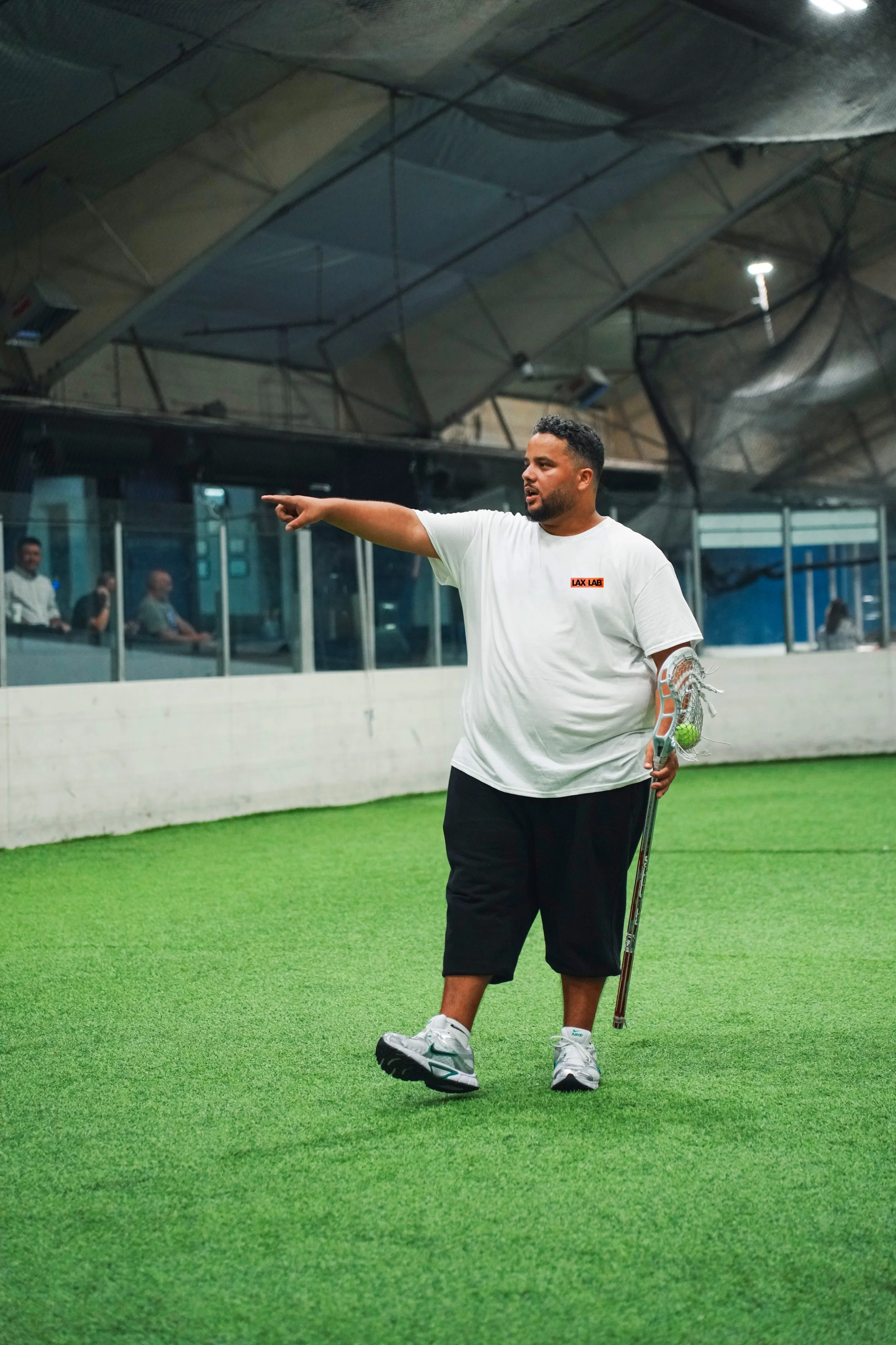 A man in a white t-shirt with a 'LAX LAB' logo on it and black shorts holding a lacrosse stick with a green ball, standing on an indoor green turf field and pointing to his left. Behind him, there are people inside a glass enclosure.