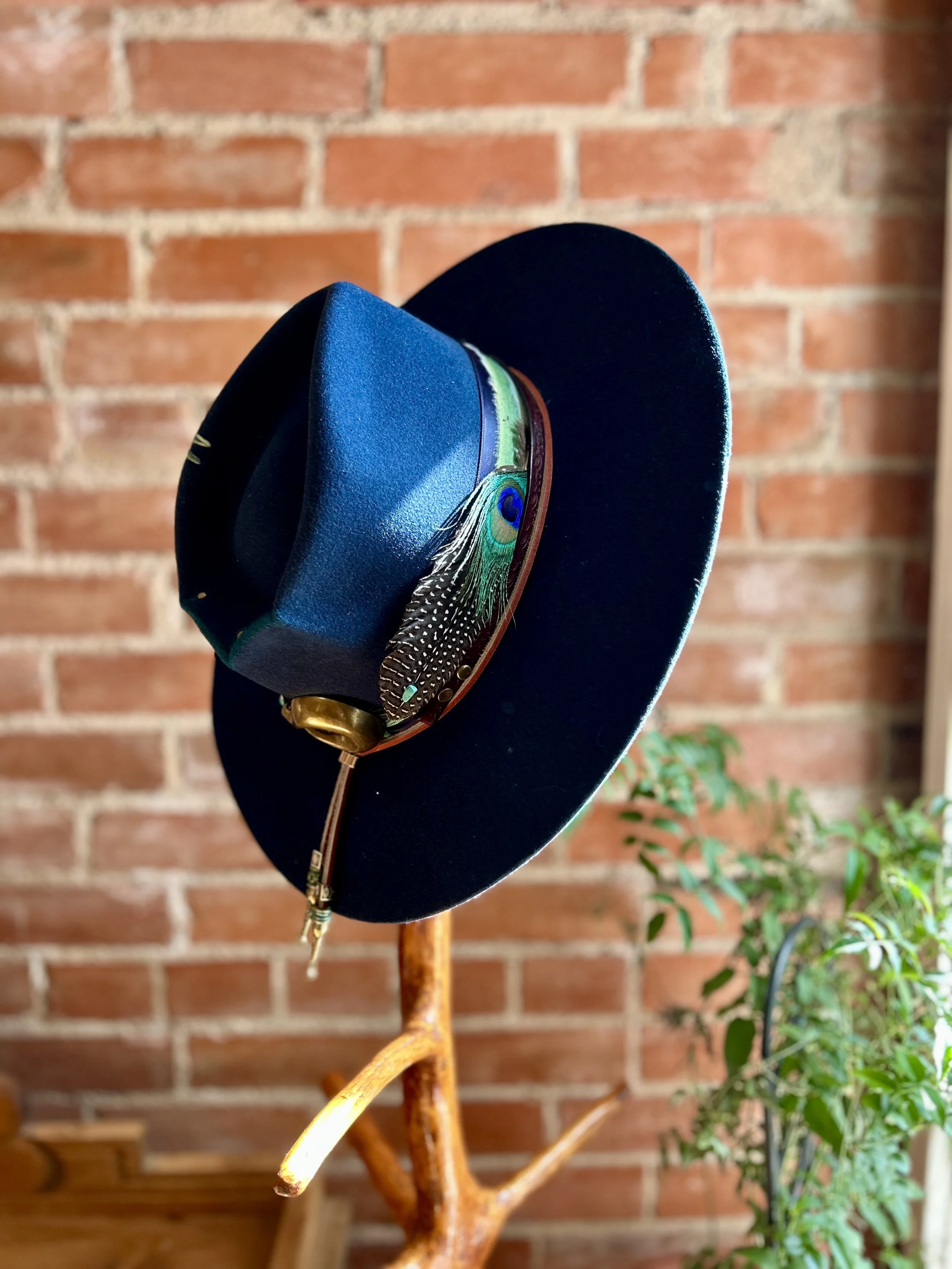 A black hat with a peacock feather on the front, hung on a wooden stand against a brick wall background in boulder colorado
