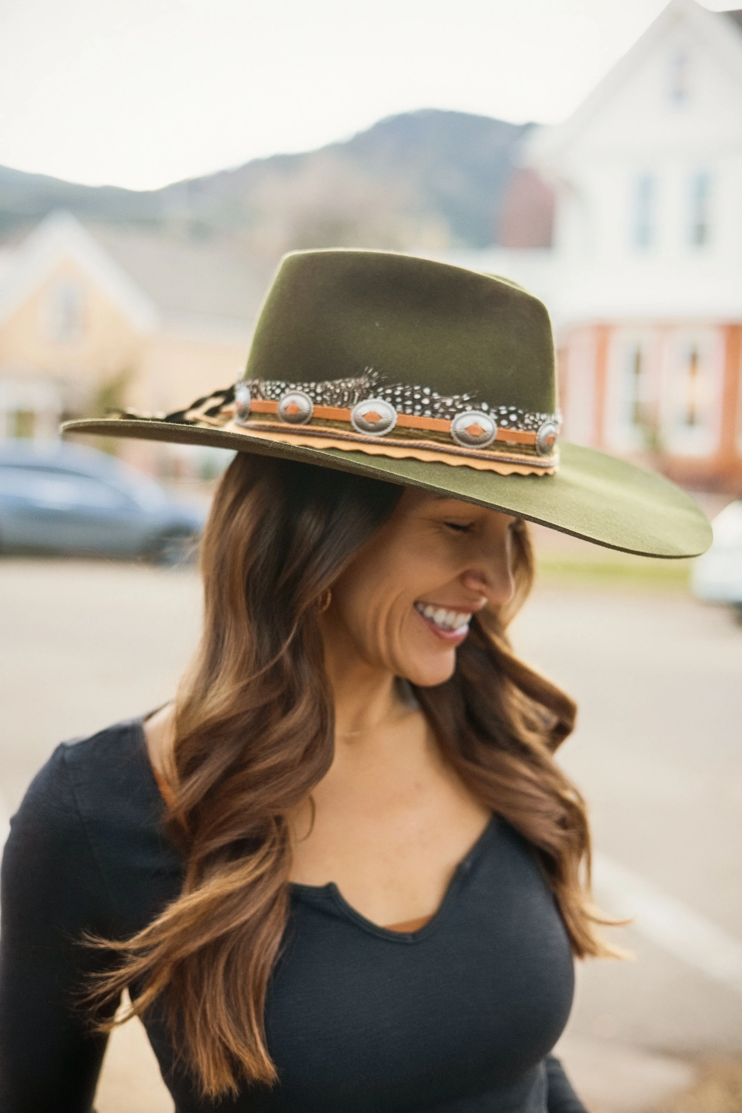 rebecca the owner of boulder brim with long, wavy brown hair smiling while wearing a dark blue top and a green hat custom decorated with buttons, feathers, and trimming.
