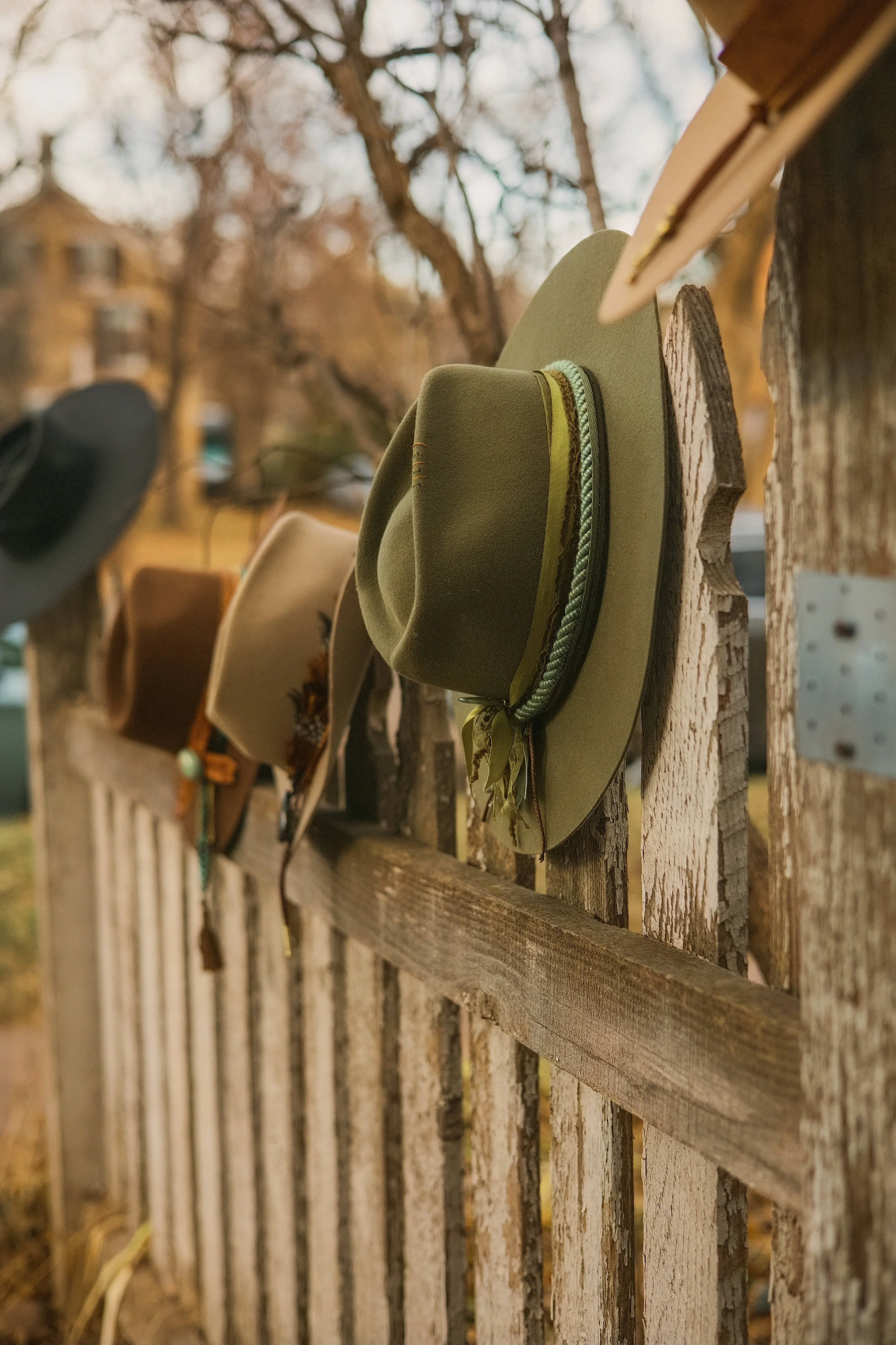 A rustic wooden fence with hats hanging on it, with blurry trees and houses in the background during autumn.