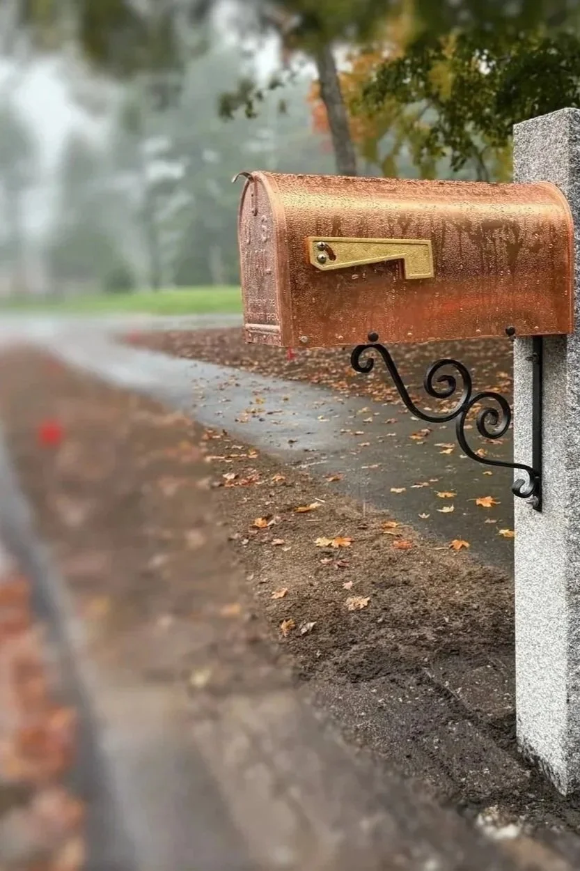 A copper mailbox mounted on a decorative black iron post on a sidewalk, with a background of autumn trees and fallen leaves