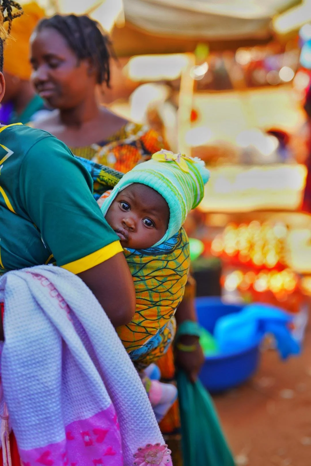 A young child dressed in a striped green hat and colorful clothing is being carried on an adult's back, looking directly at the camera with wide eyes. The background features a busy marketplace with blurred figures and vibrant stalls.