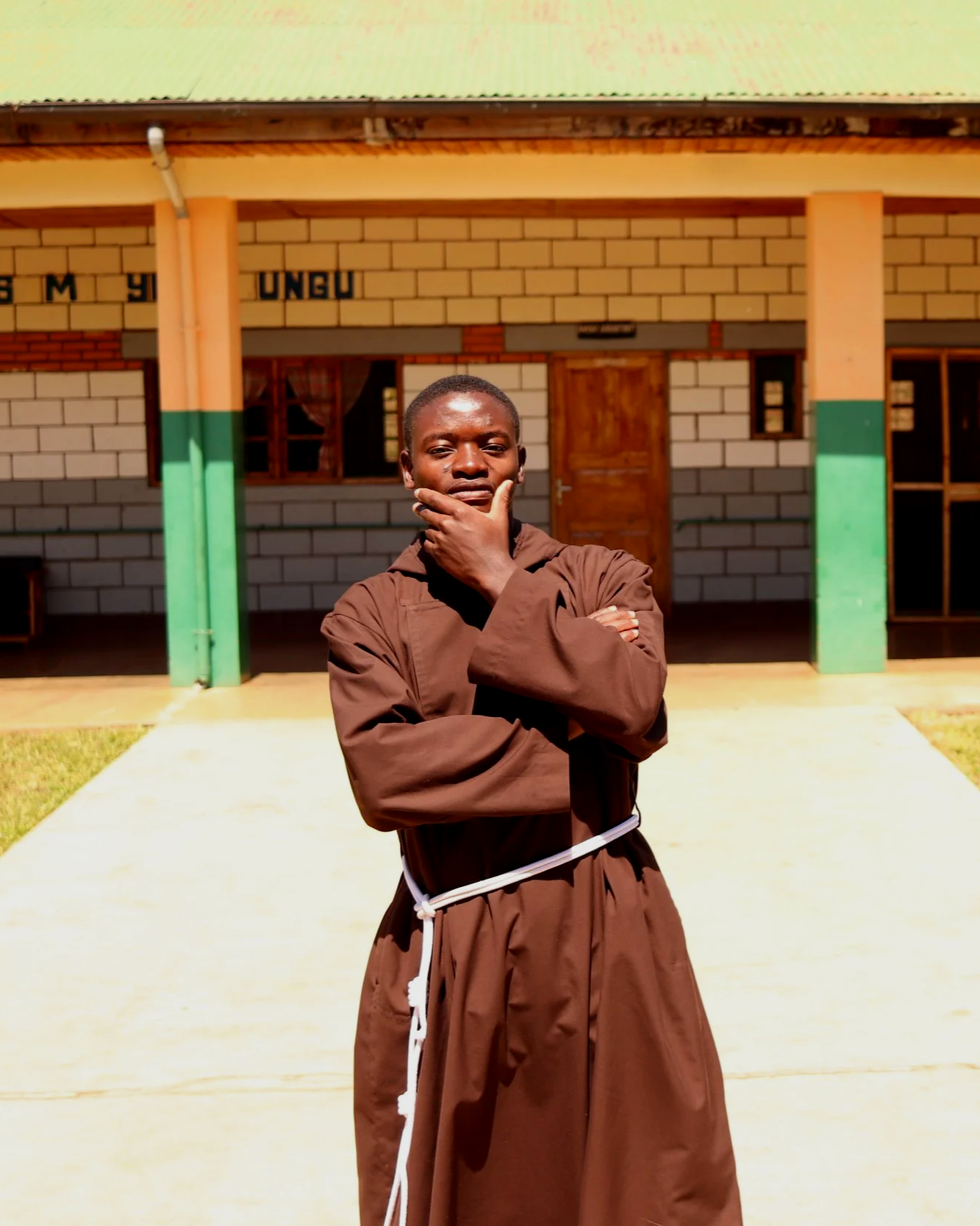 A young man in brown religious robes standing outside a building with a thoughtful expression, hand on his chin.