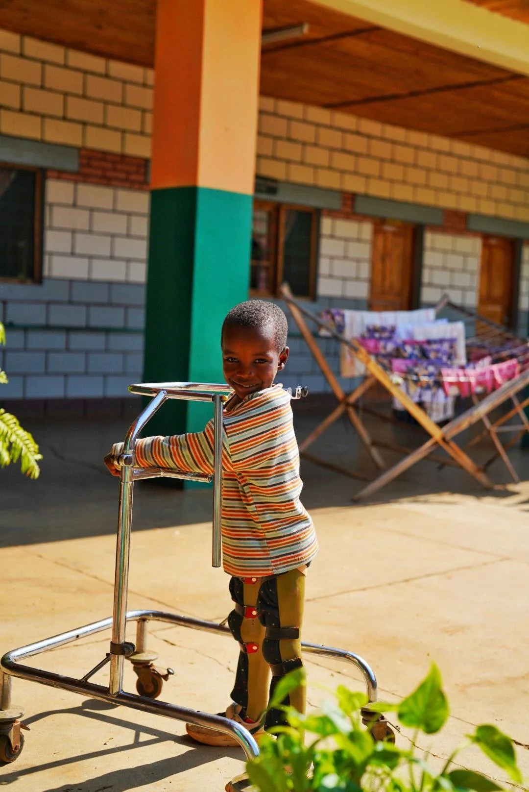A young boy smiling while standing with a walker outside a building with a brick wall and colorful laundry hanging on a drying rack in the background.