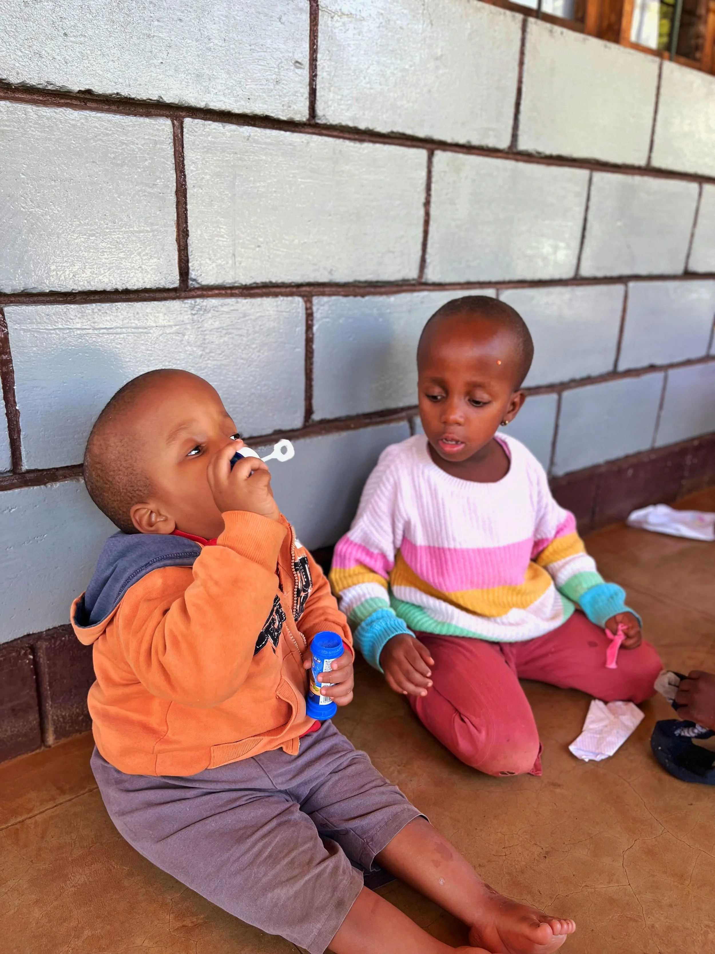 Two young children sitting on the floor against a brick wall. The boy on the left is holding a toy or object to his mouth, and the girl on the right is watching him. The girl is wearing a colorful striped sweater, and the boy is wearing an orange hoodie.