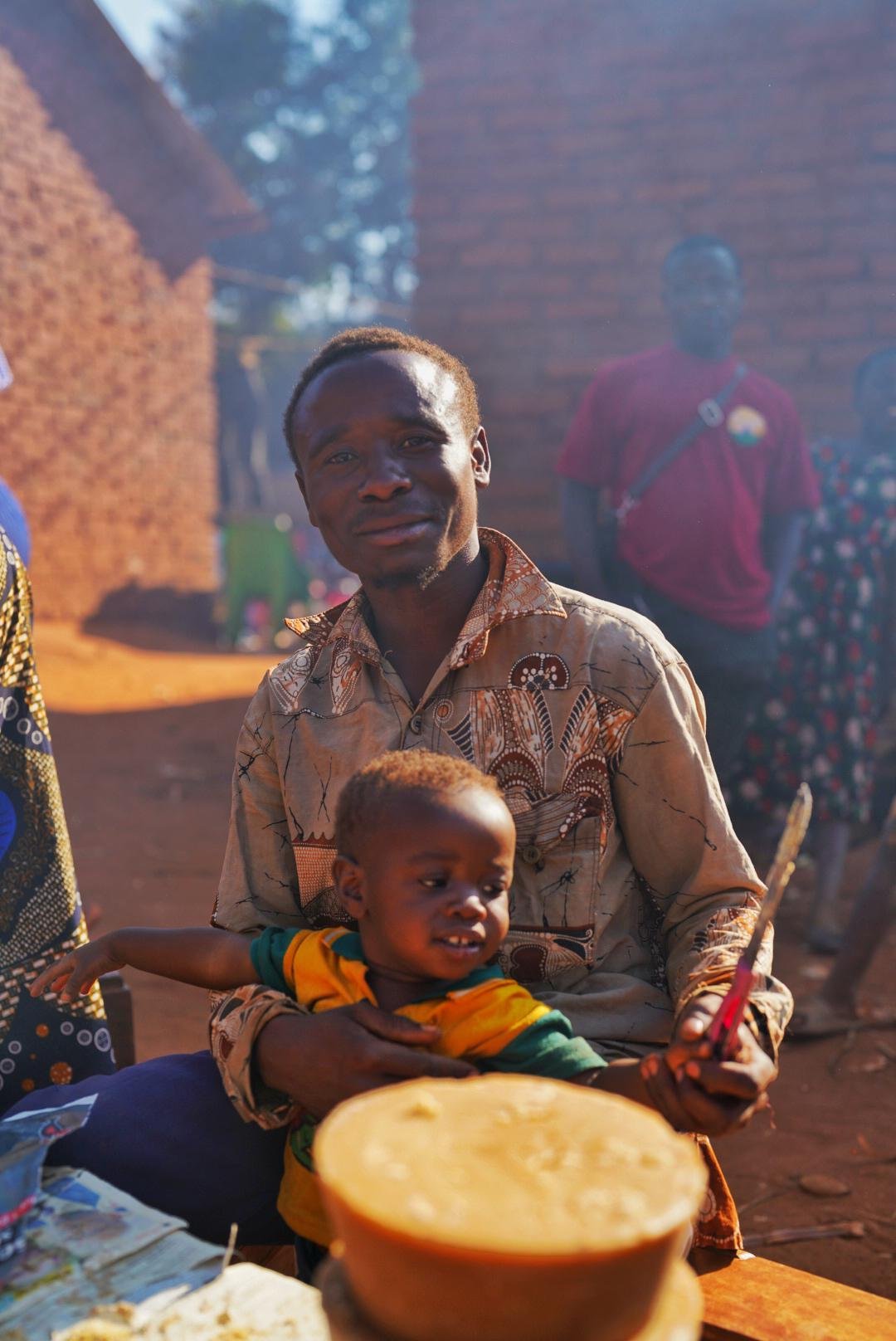A man holding a young child in a rural village setting, with several people and brick structures in the background.