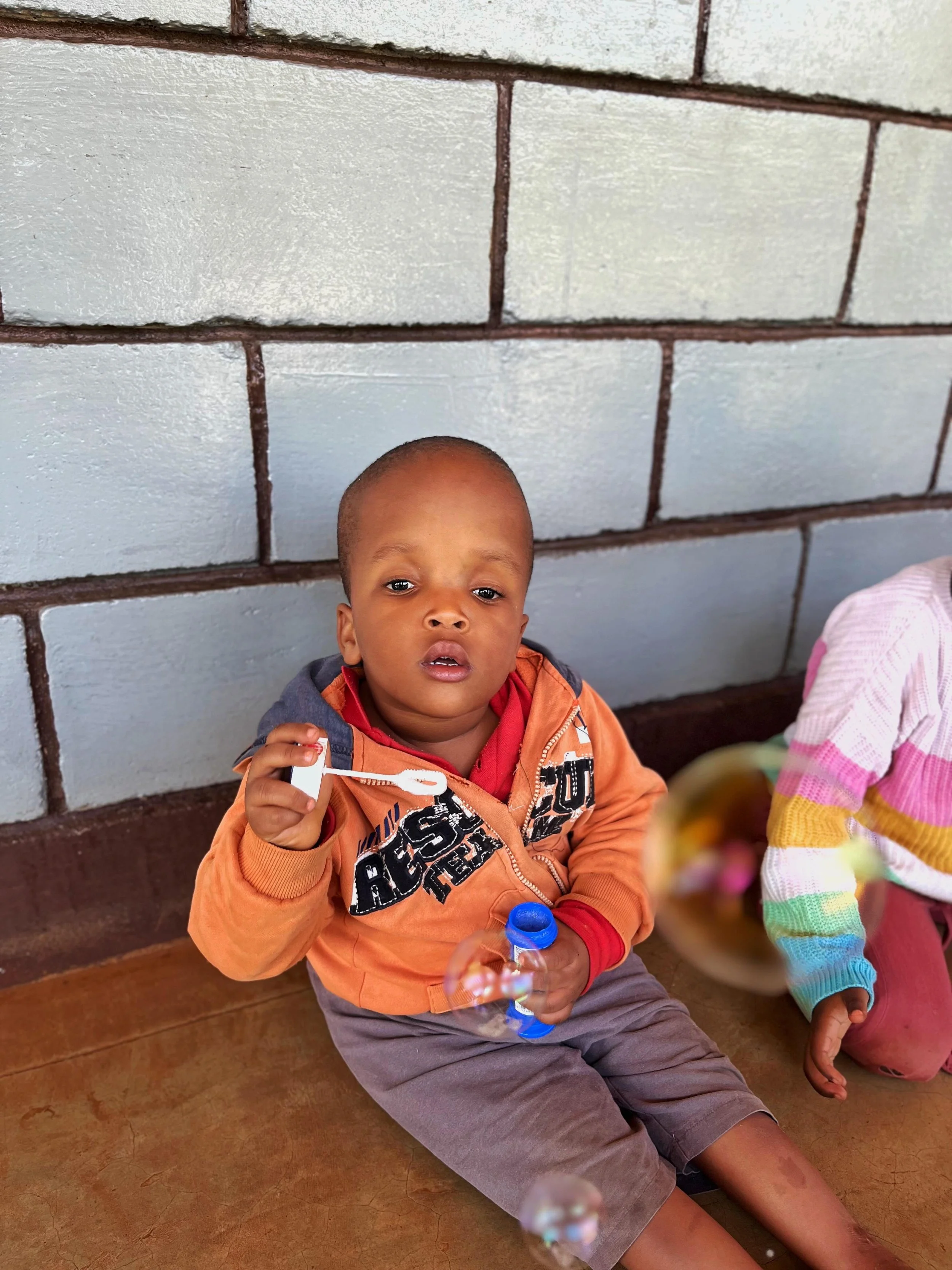 A young boy sitting on a wooden floor against a gray brick wall, holding a bubble wand and a bubble solution container, with soap bubbles floating around him.