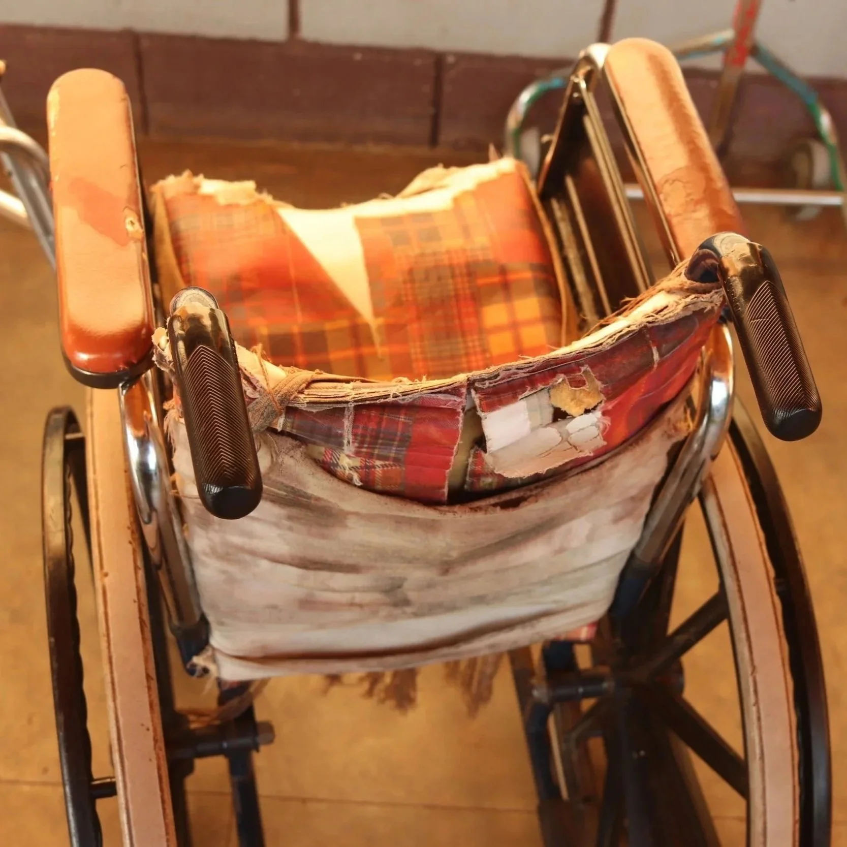 An old, worn-out wheelchair with a ripped, plaid fabric seat and backrest, showing signs of heavy use and damage, with parts of the fabric peeling off.