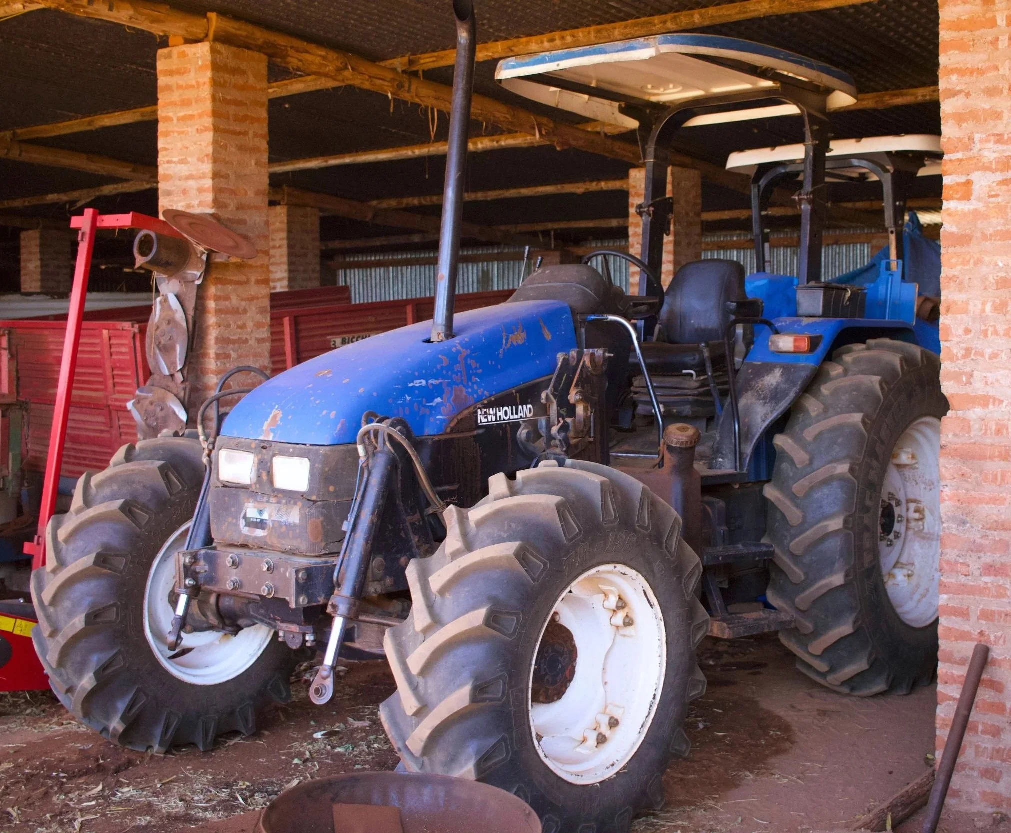 A blue New Holland tractor with large white wheels inside a brick and wood shed, surrounded by farm equipment and tools.