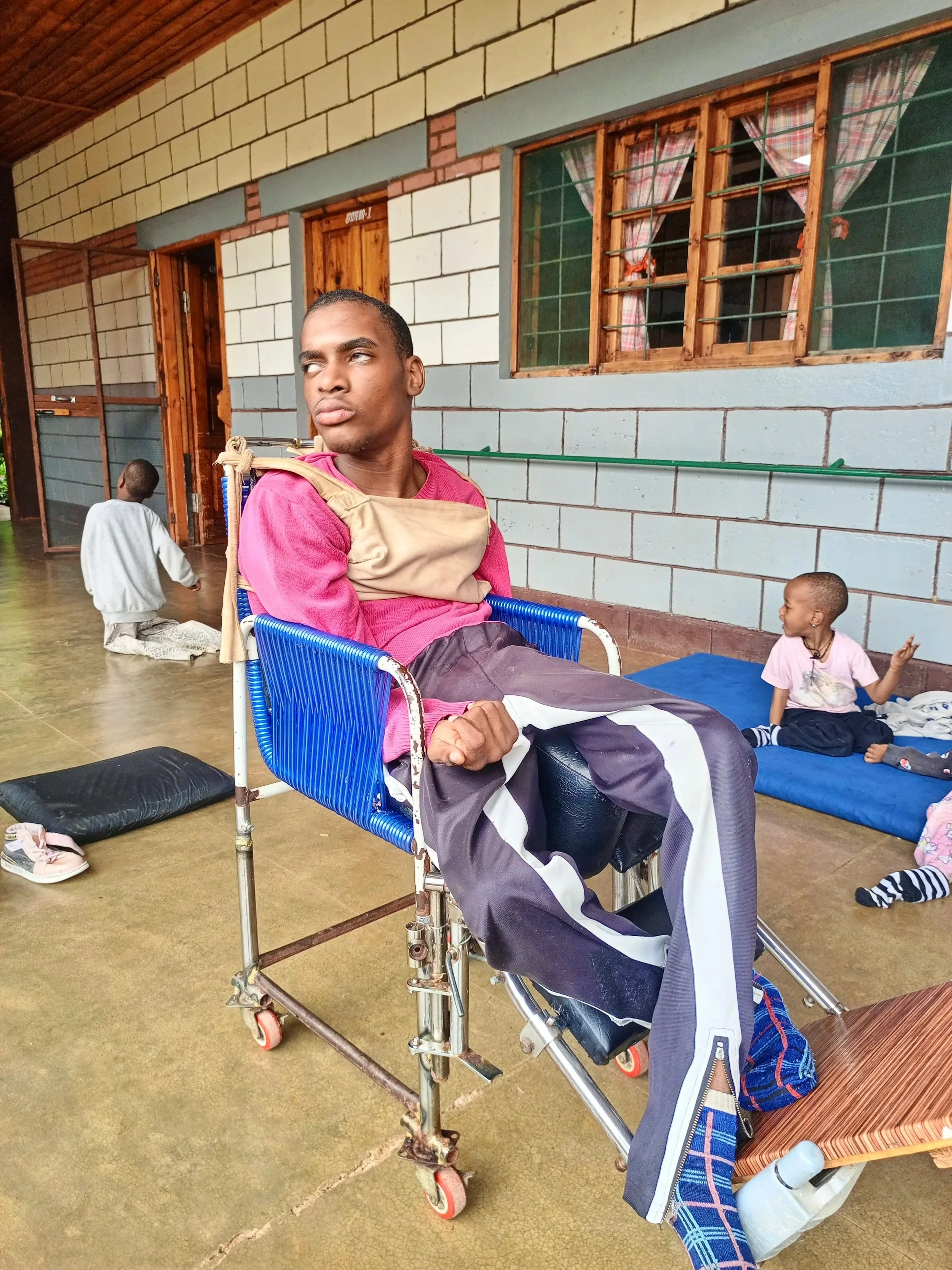 A young man with a serious expression sitting in a blue wheelchair inside a room with a concrete floor and stone wall. Two children are sitting on a mat in the background, one facing sideways and the other sitting with their back to the camera.