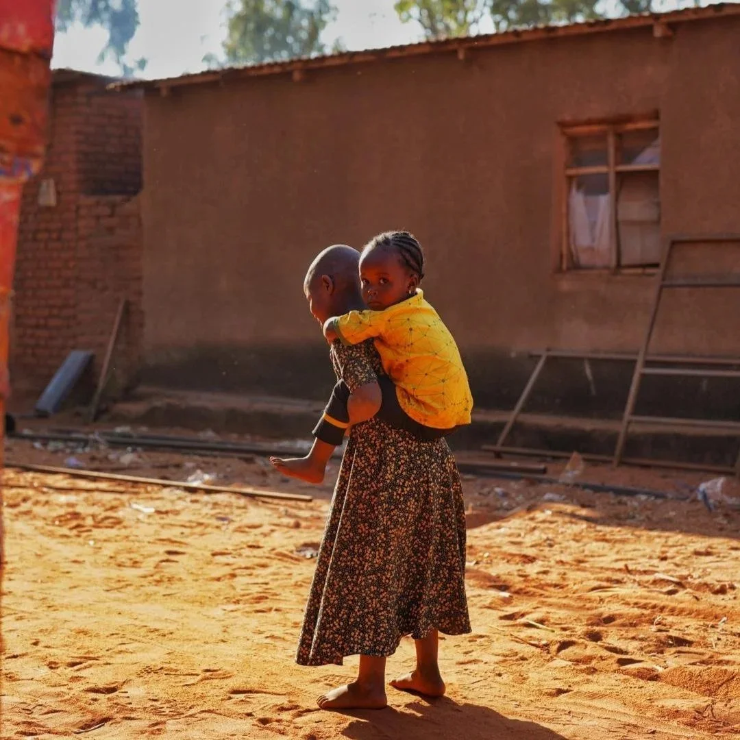A woman carrying a child on her back walking outdoors on a dirt ground in a rural area, with a simple house in the background.