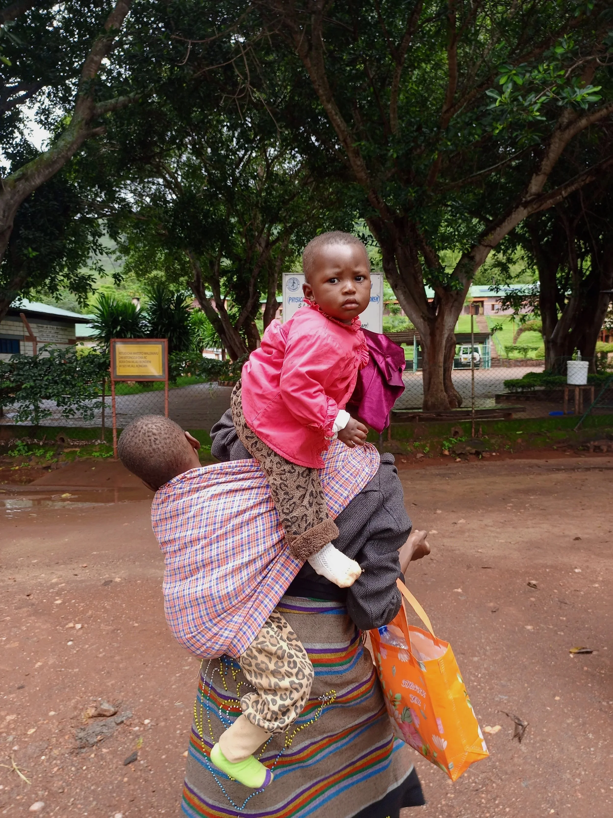 A woman carrying a young girl on her back outdoors. The girl is wearing a pink jacket, leopard print pants, and white socks, and is looking at the camera with a serious expression. The background features trees and a fence.