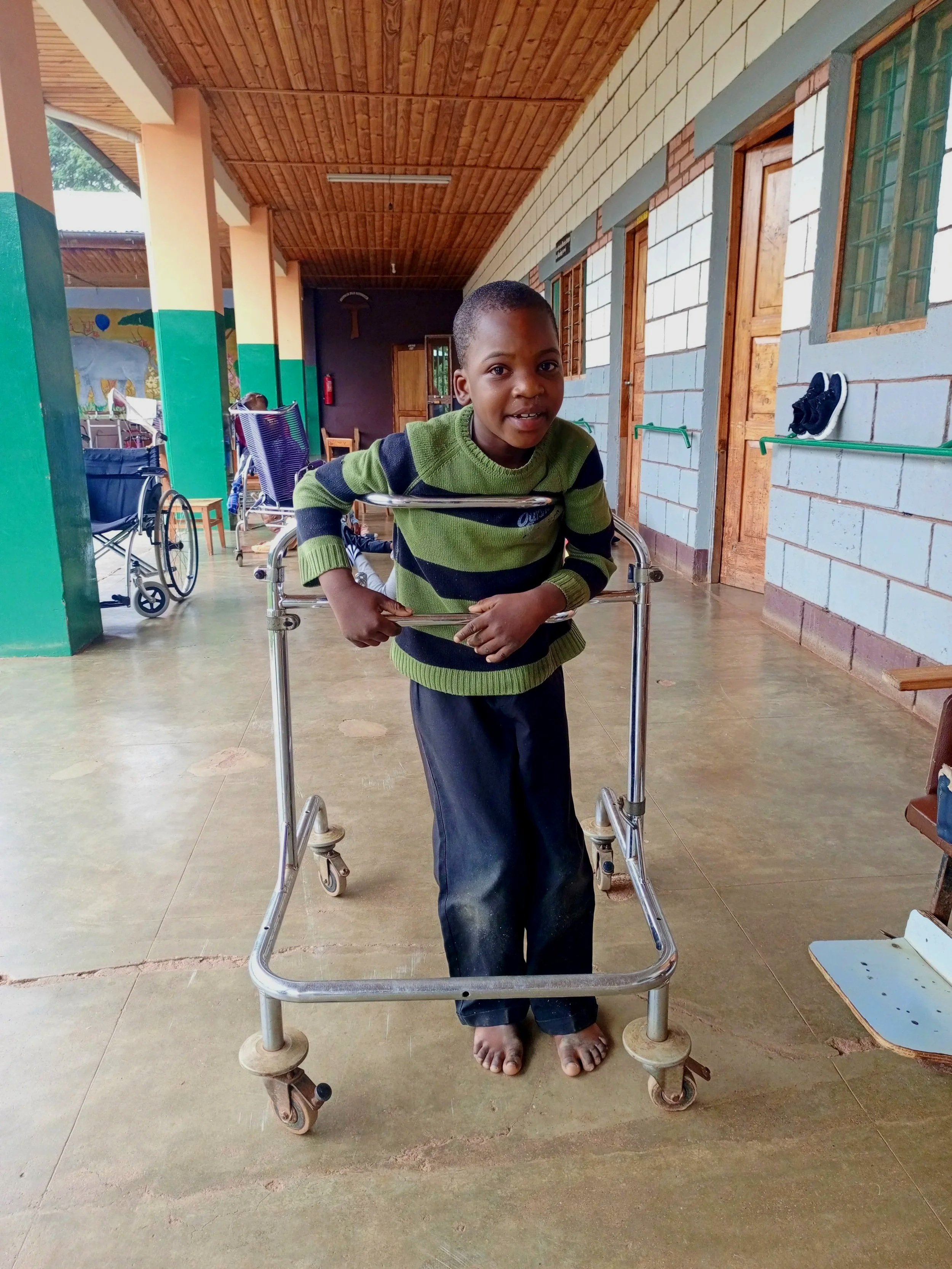 A young boy standing with the help of a walker in a room with a wooden ceiling and brick walls, multiple wheelchairs in the background.