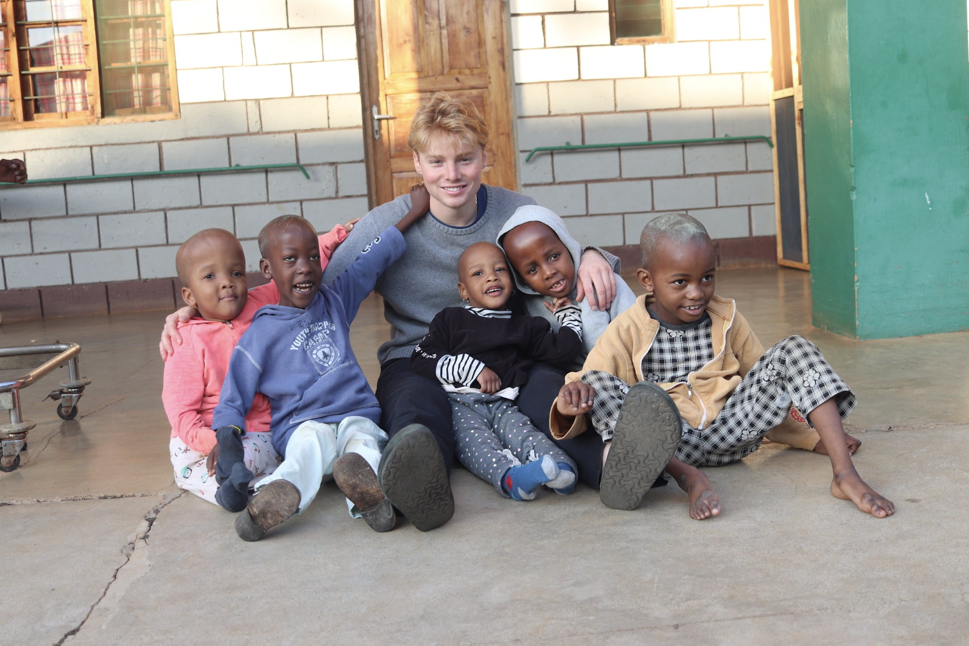 A young man sitting on the floor with five children in a room with concrete flooring and brick walls.