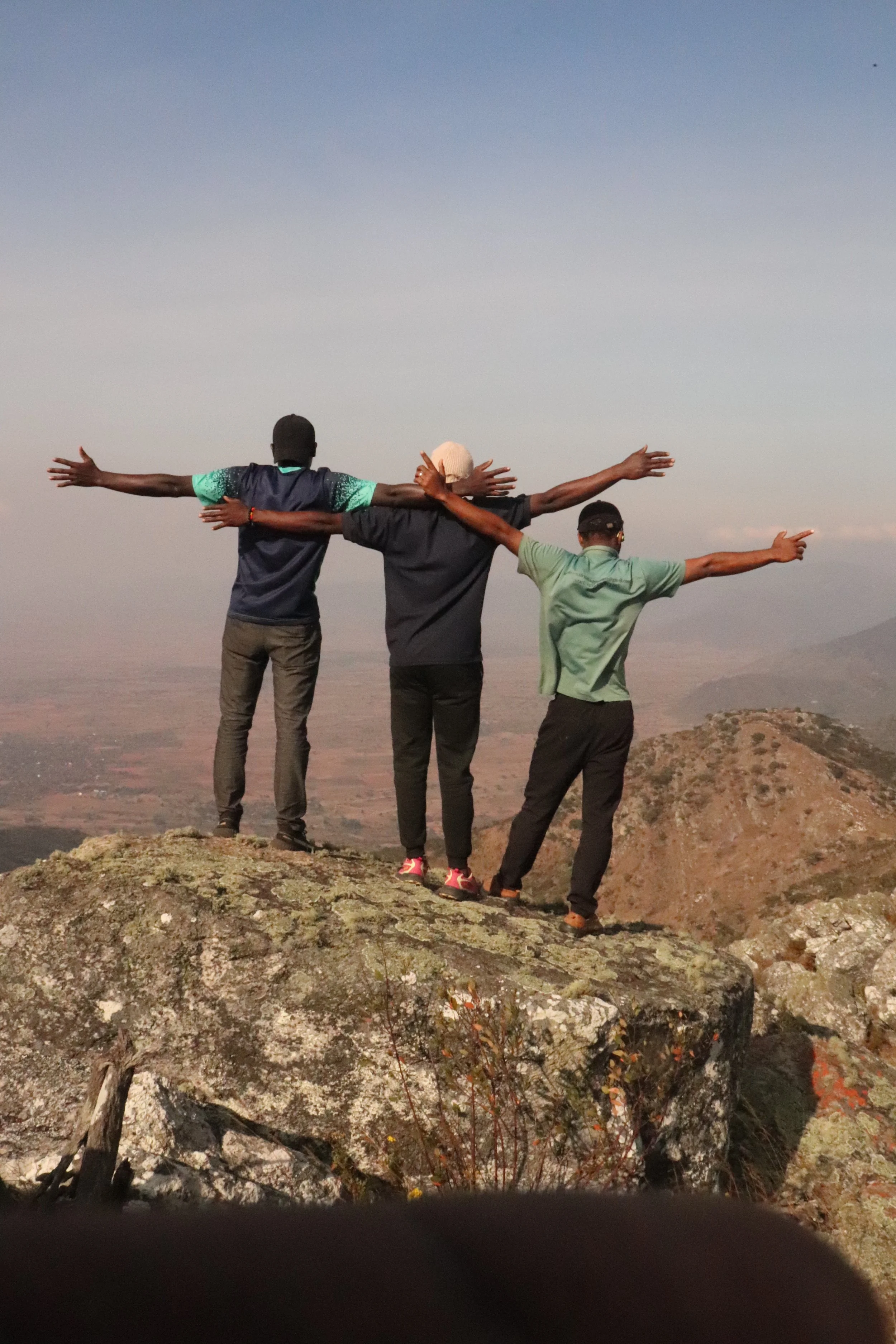 Three people standing on a mountain top with arms outstretched, overlooking a vast landscape and mountains in the distance during daytime.