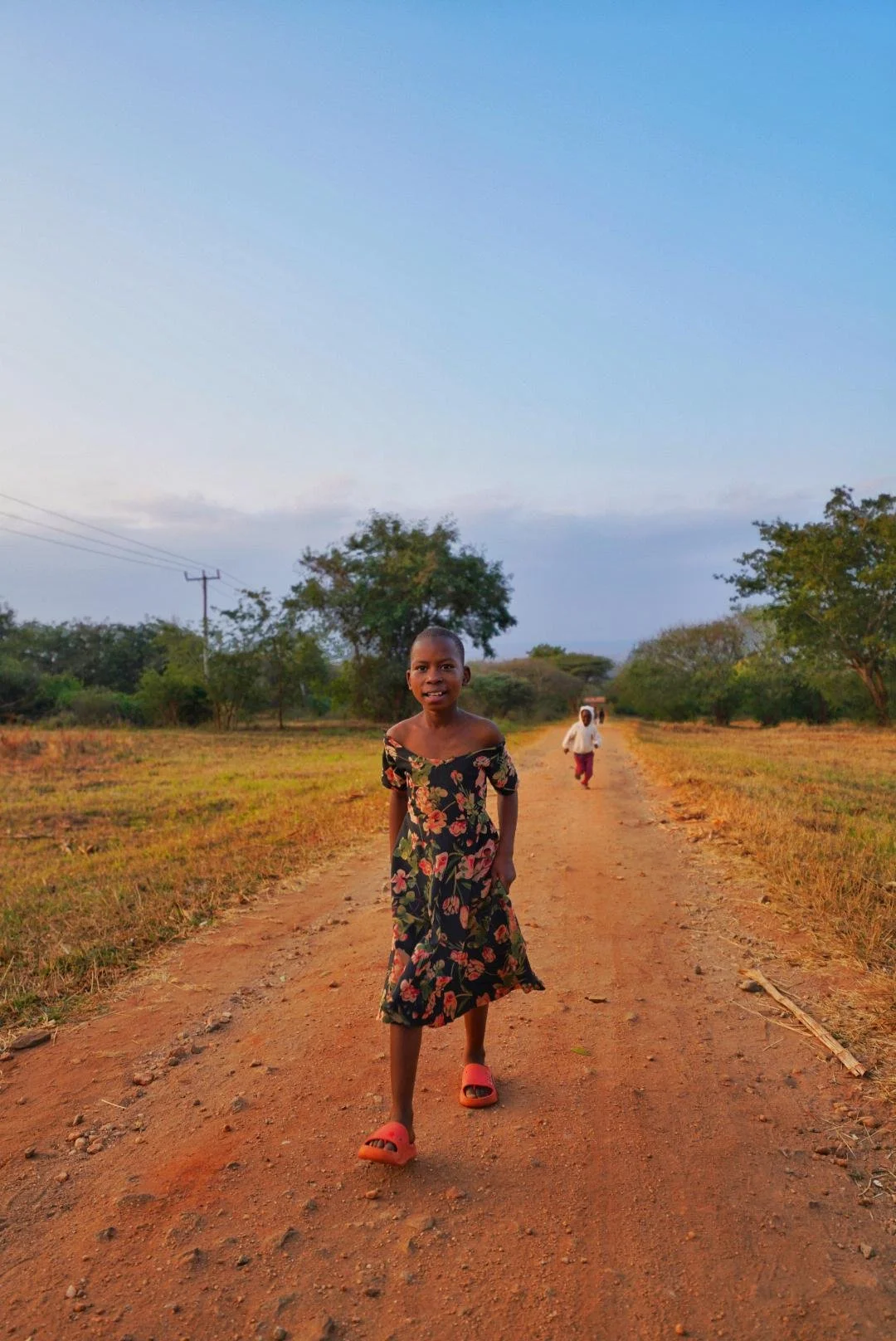 A young girl walking on a dirt path in a rural area, wearing a floral dress and orange sandals, with two other people in the background and trees on either side of the path under a blue sky.