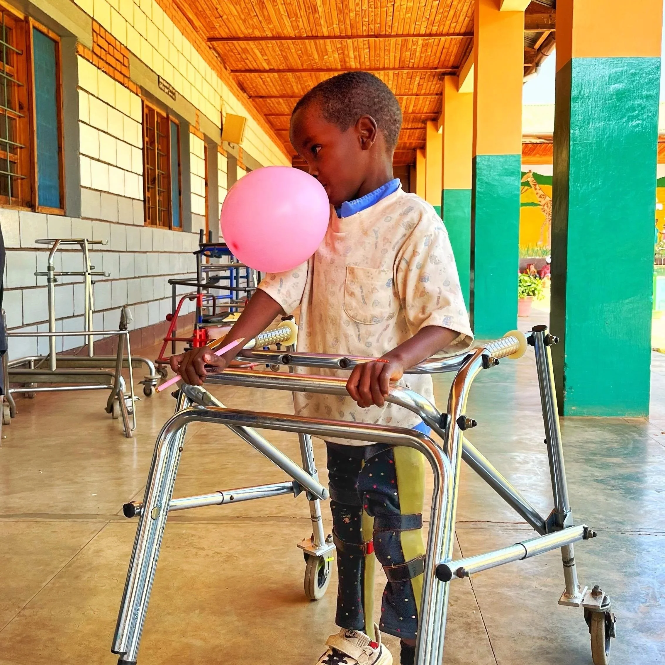 A young boy in a hospital gown using a walker, holding a pink balloon and looking at it thoughtfully in a covered outdoor area with colorful columns and walls.