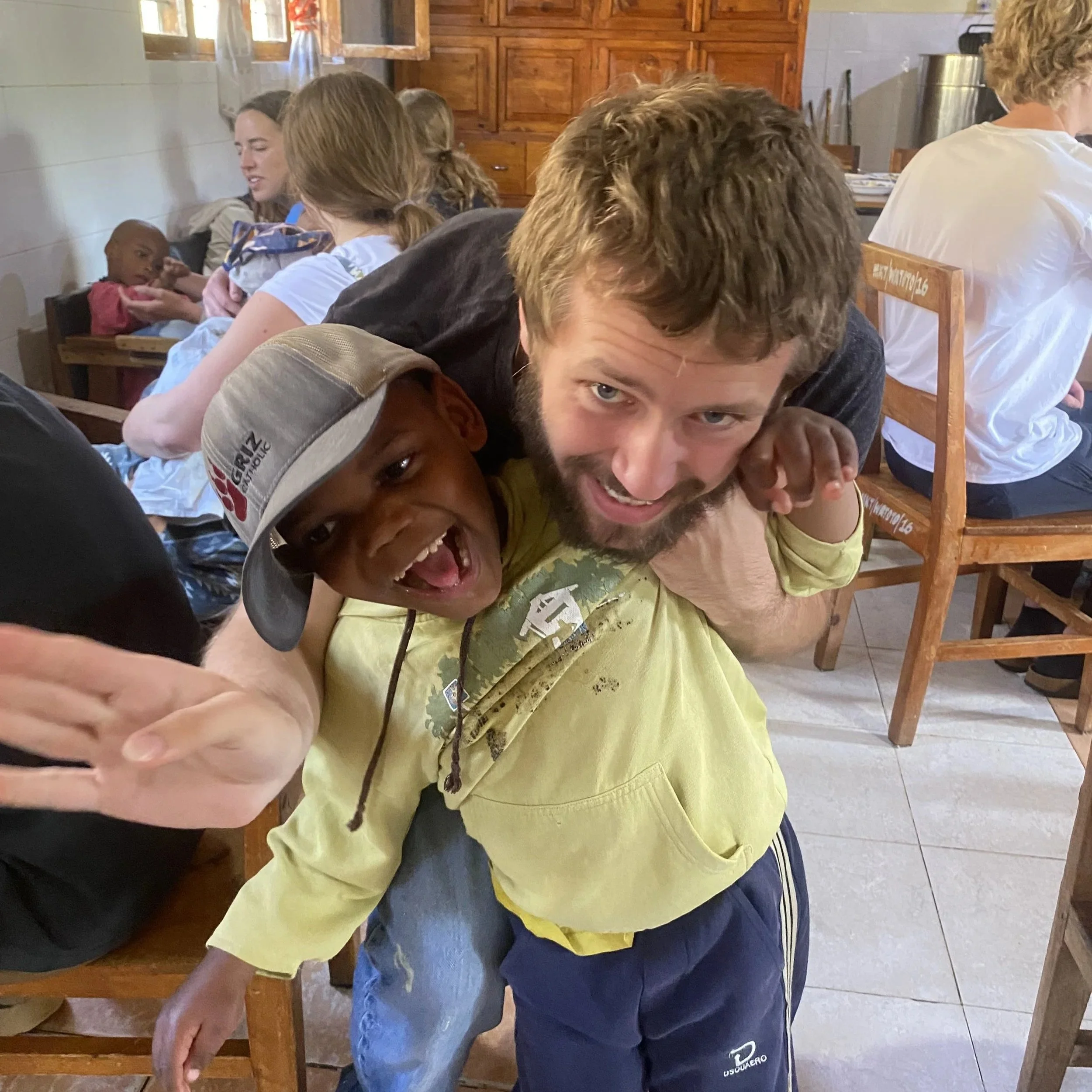 A joyful young boy and a man playfully mimic a hug in a room with other children and adults sitting and interacting in the background.