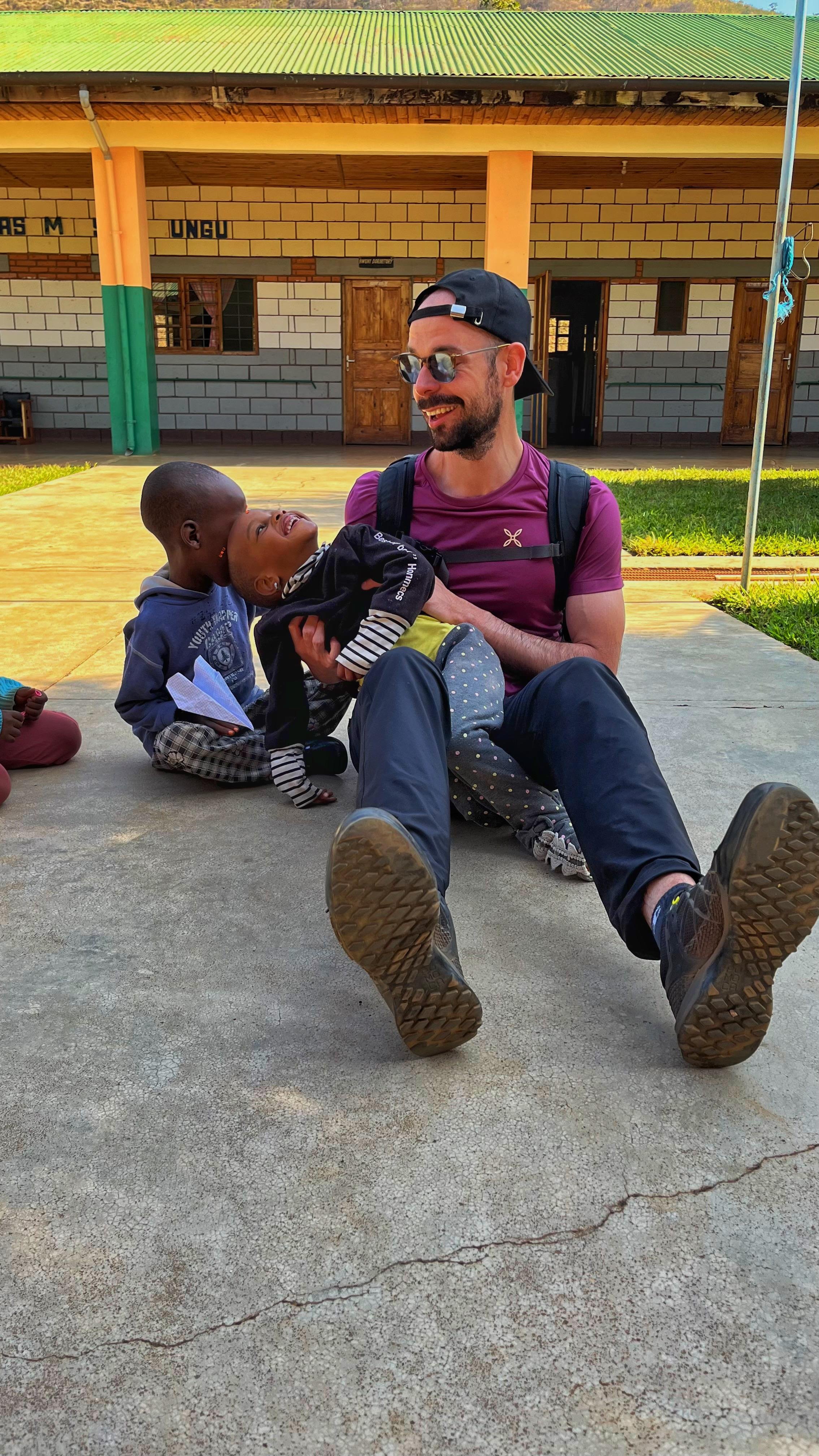 A man with a beard, wearing sunglasses, a black cap, a purple shirt, and a backpack, sitting outdoors on a concrete surface, laughing with two children, one of whom is lying on his lap, while other children sit nearby in front of a building with brick and wall tiles, under a green metal roof