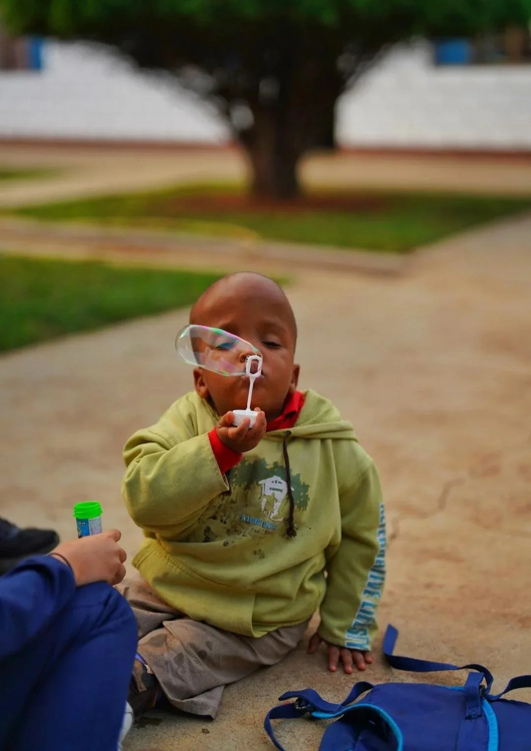 A young boy in a yellow hoodie blowing a bubble with bubblegum, sitting on the ground outdoors. There is a blue backpack nearby and another person holding a container of bubblegum bubble solution. A tree and some buildings are visible in the background.