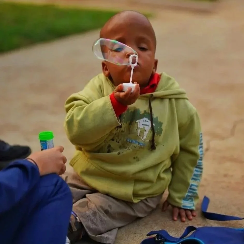 A young child sitting on the ground blowing bubbles with a bubble wand, wearing a green hoodie and beige pants.