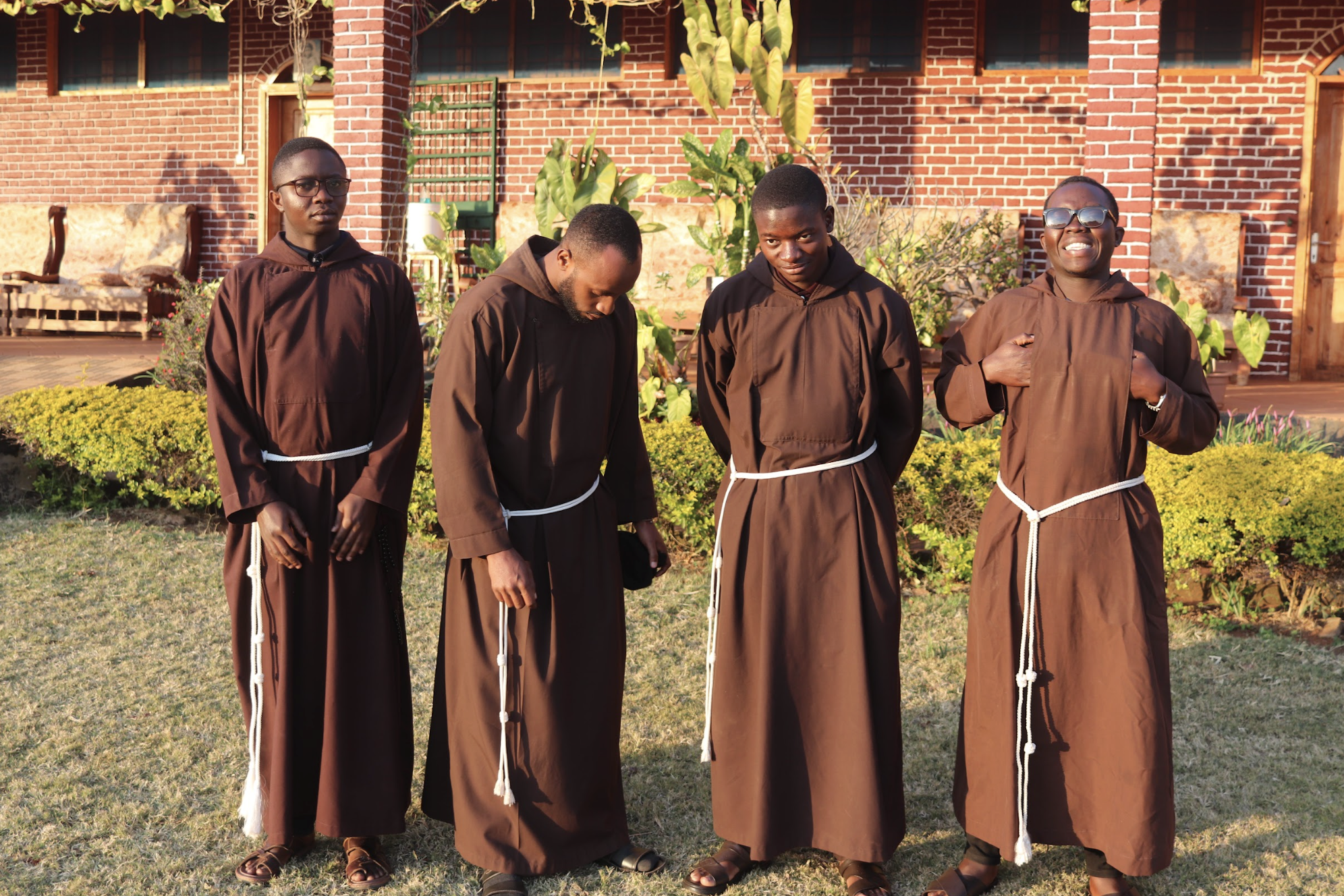 Four young men dressed in brown religious robes with white ropes, standing outdoors in front of a brick building and plants, during daytime.