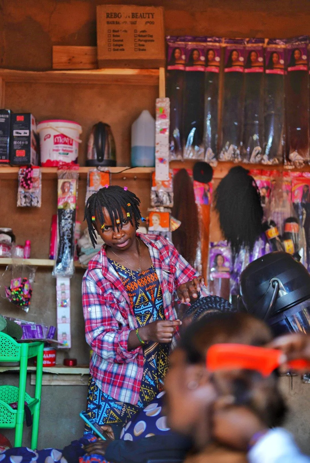 A woman with braided hair decorated with colorful beads, wearing a plaid shirt over a patterned dress, working behind the counter of a small shop. The shop has various hair products and accessories on shelves and hanging on the wall.