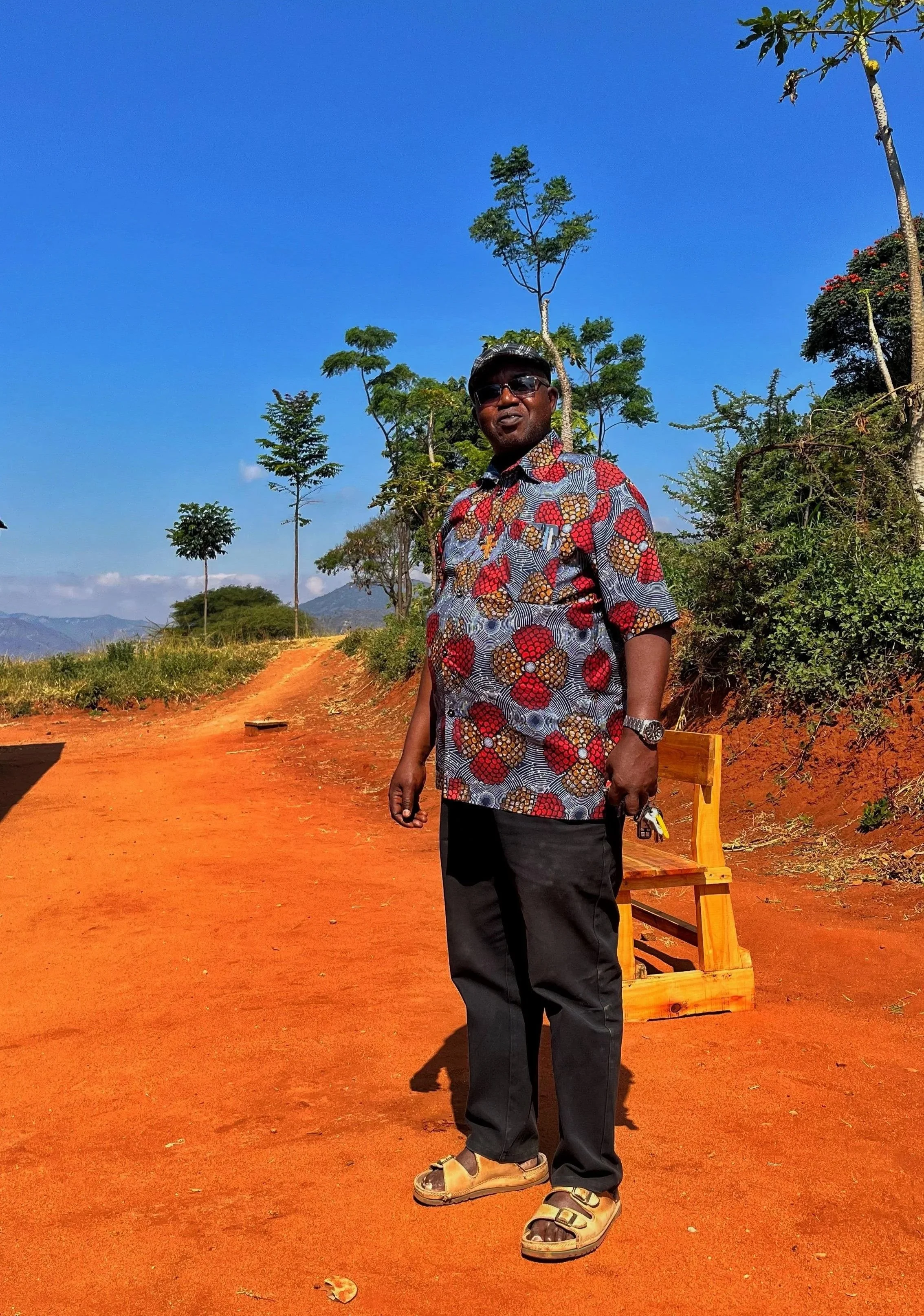 A man wearing sunglasses, a colorful patterned shirt, black pants, and sandals standing outdoors on an orange dirt path surrounded by sparse trees and a bright blue sky.