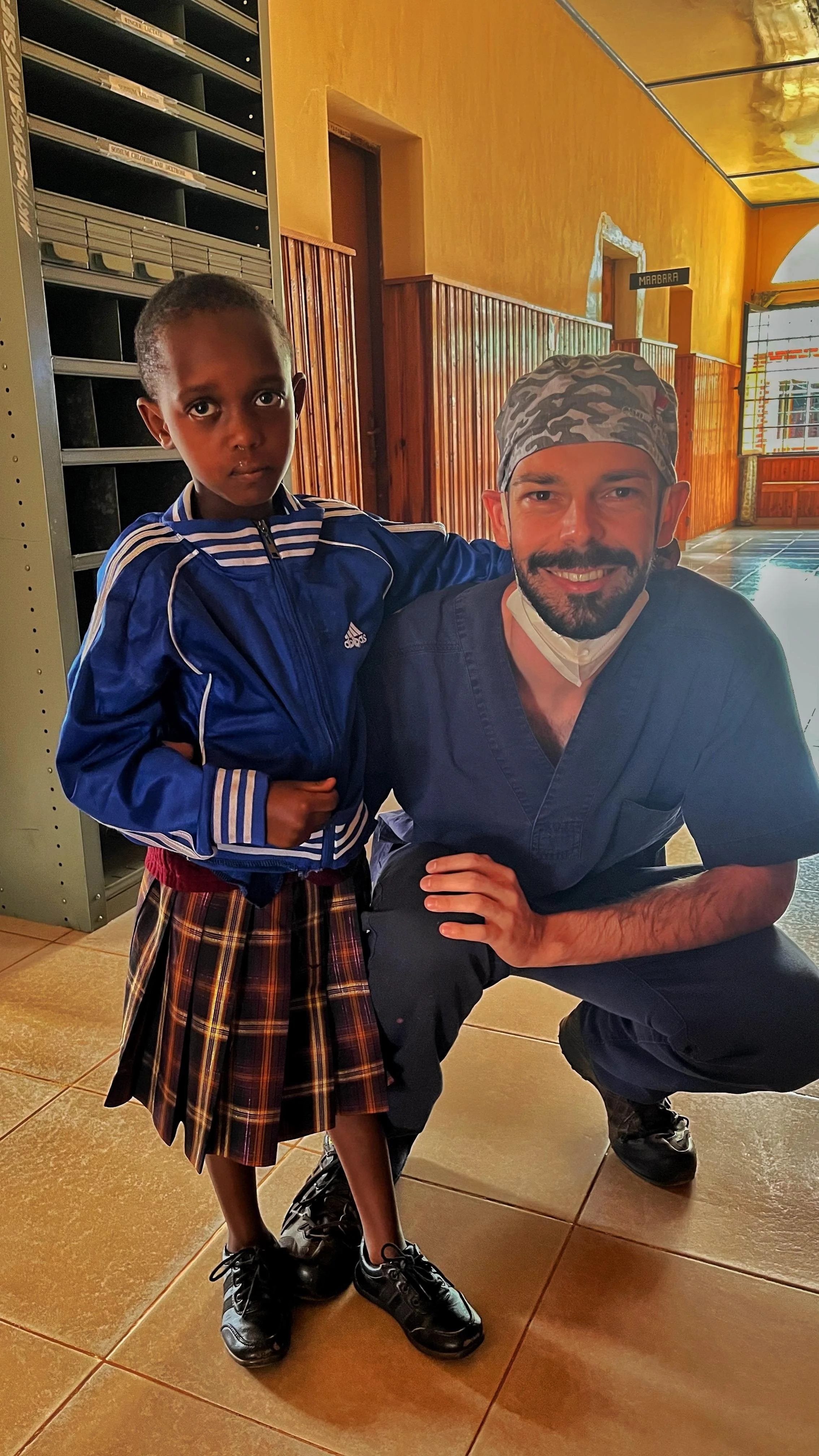 A young girl in a plaid skirt and blue jacket standing next to a man in medical scrubs inside a building with wooden paneling and a yellow wall.