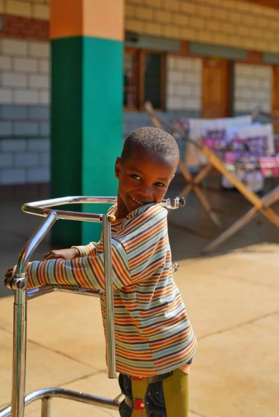 A young boy with a short haircut and wearing a striped long-sleeve shirt stands outdoors, smiling and leaning on a metal walker in front of a background featuring a brick building and laundry hanging on a clothesline.