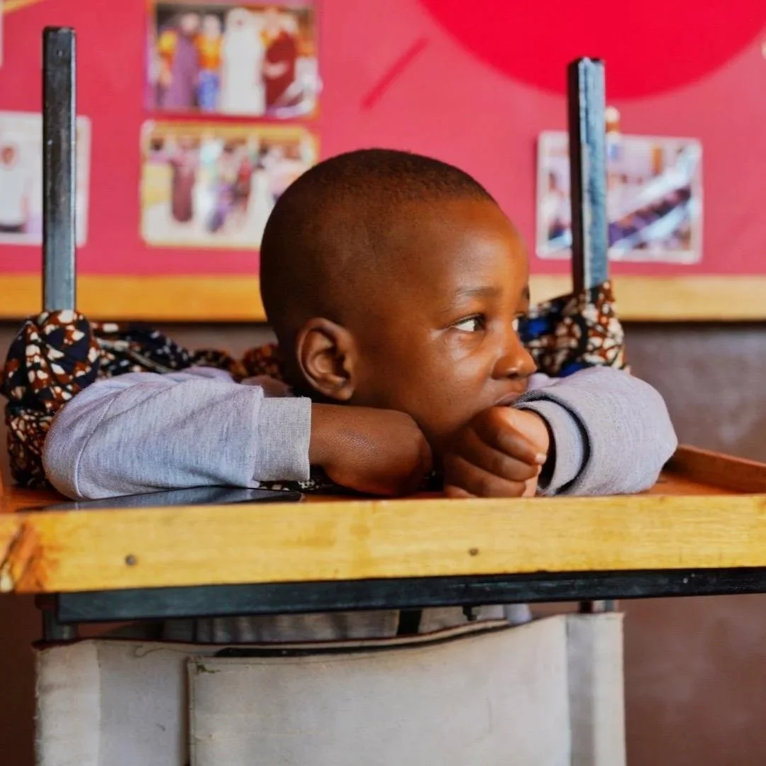 A young boy with a short haircut resting his head on a desk in a classroom, with colorful pictures on a pink wall in the background.