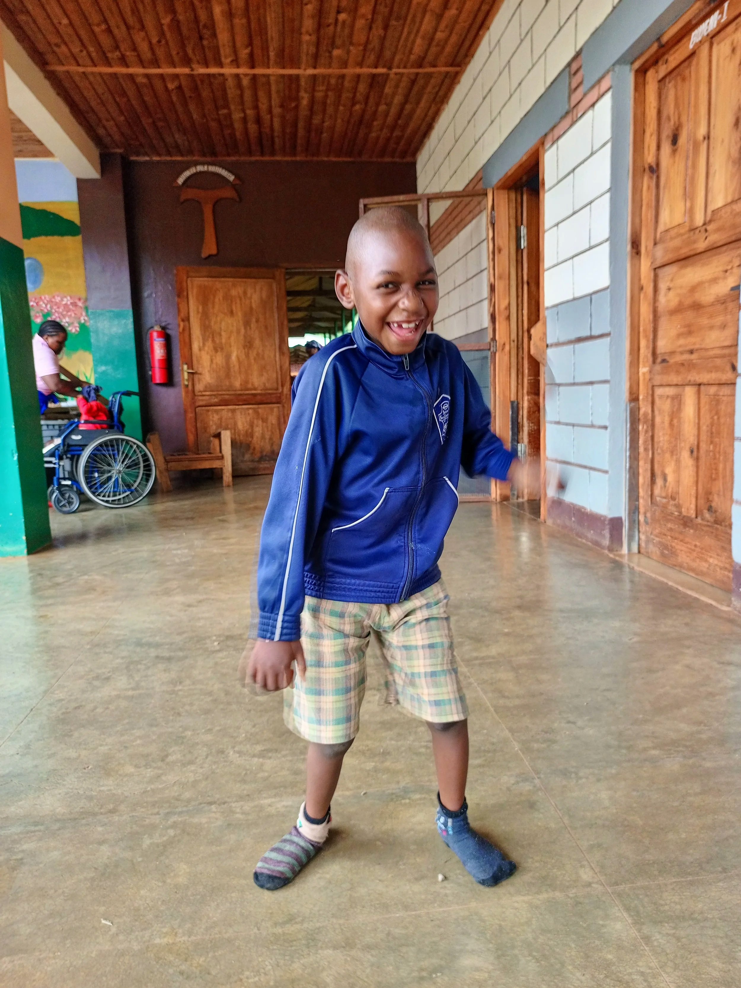 A young boy with a shaved head smiling and sticking out his tongue, wearing a blue jacket, plaid shorts, and mismatched socks, standing indoors with wooden doors and colorful walls in the background.