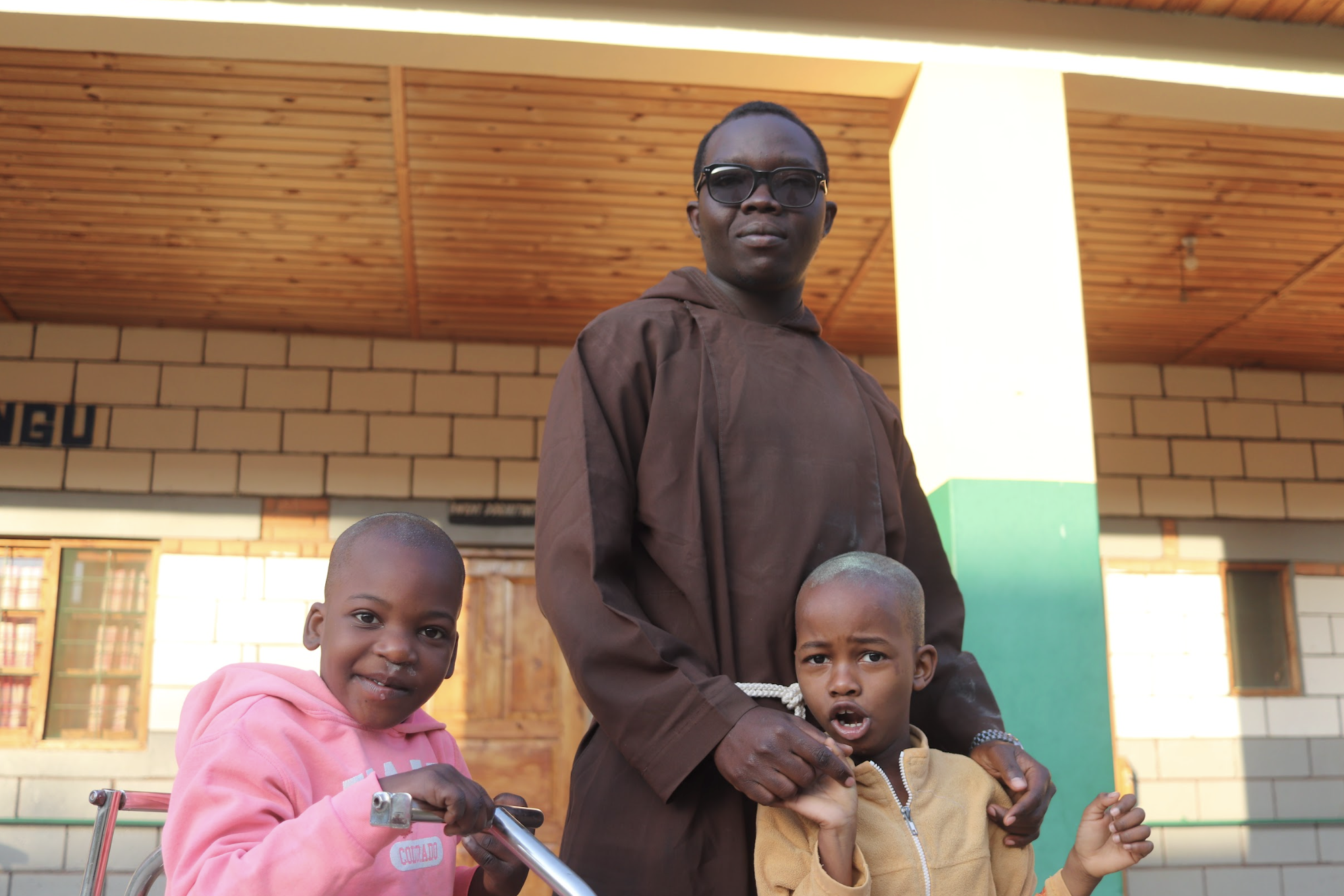 A man with glasses standing outdoors with two young children, one girl in a pink hoodie and one boy in a tan jacket, in front of a building with a brick wall and wooden ceiling.