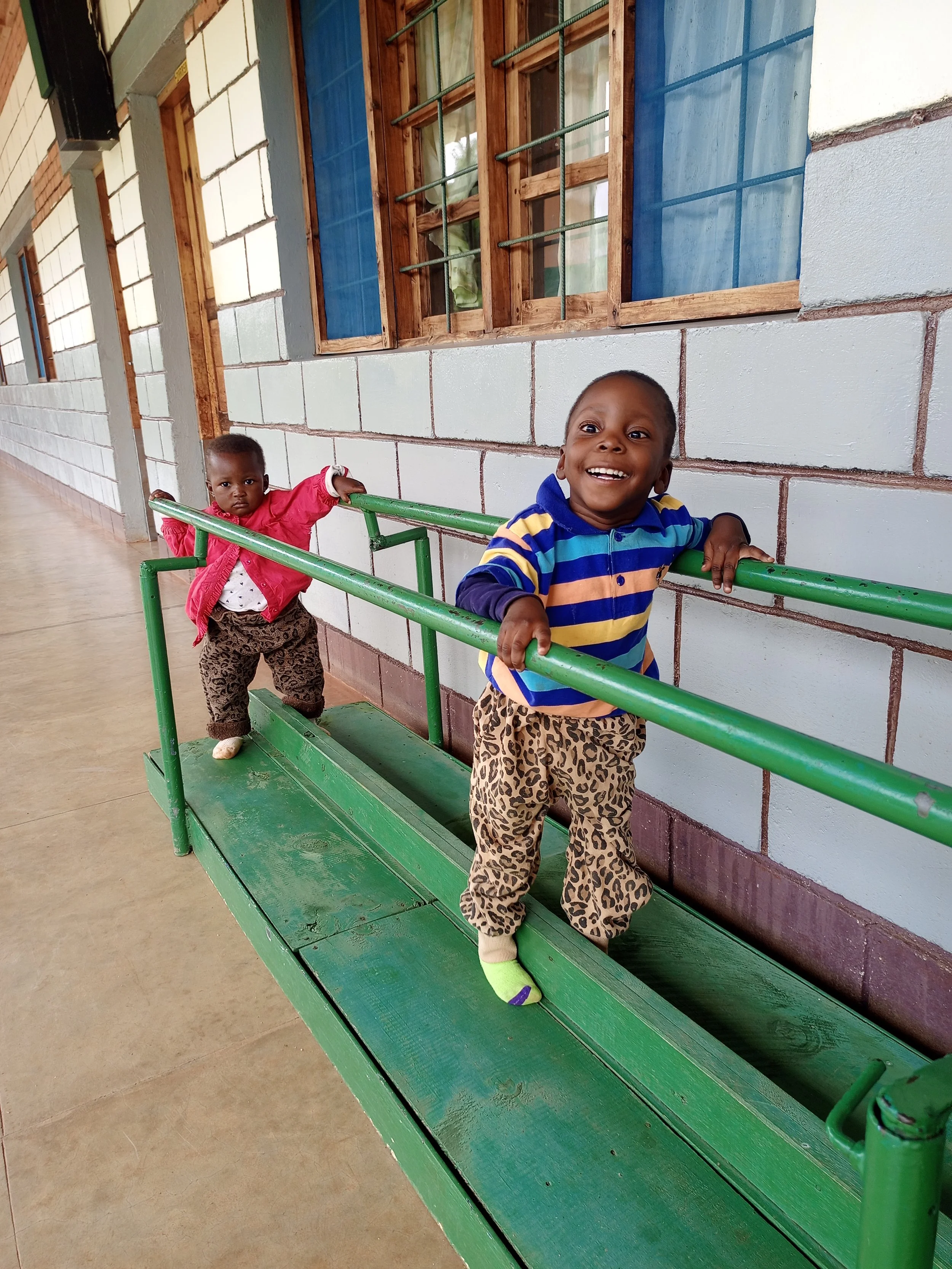 Two young children playing on a green outdoor bench with railing, standing against a brick wall outside a building with windows.