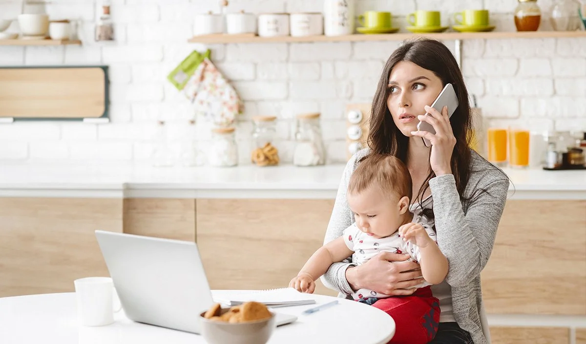 Woman on a phone holding a baby in a kitchen with a laptop, mug, and bowl of cookies on the table.