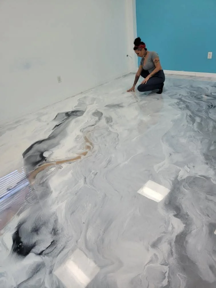 Woman kneeling on a marble-like epoxy resin floor, working on the surface in a room with white and blue walls.