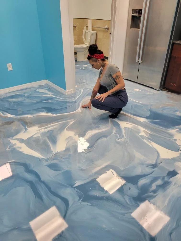 Woman in gray shirt and navy pants kneeling on a resin floor with swirling blue and white marble-like epoxy flooring.