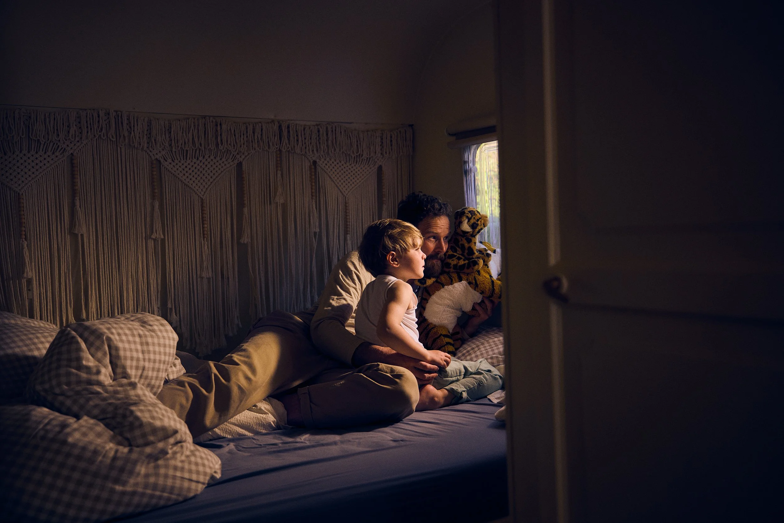 Ein Mann und ein Kind sitzen abends in einem Bett, schauen durch ein Fenster und spielen mit einem Teddybär