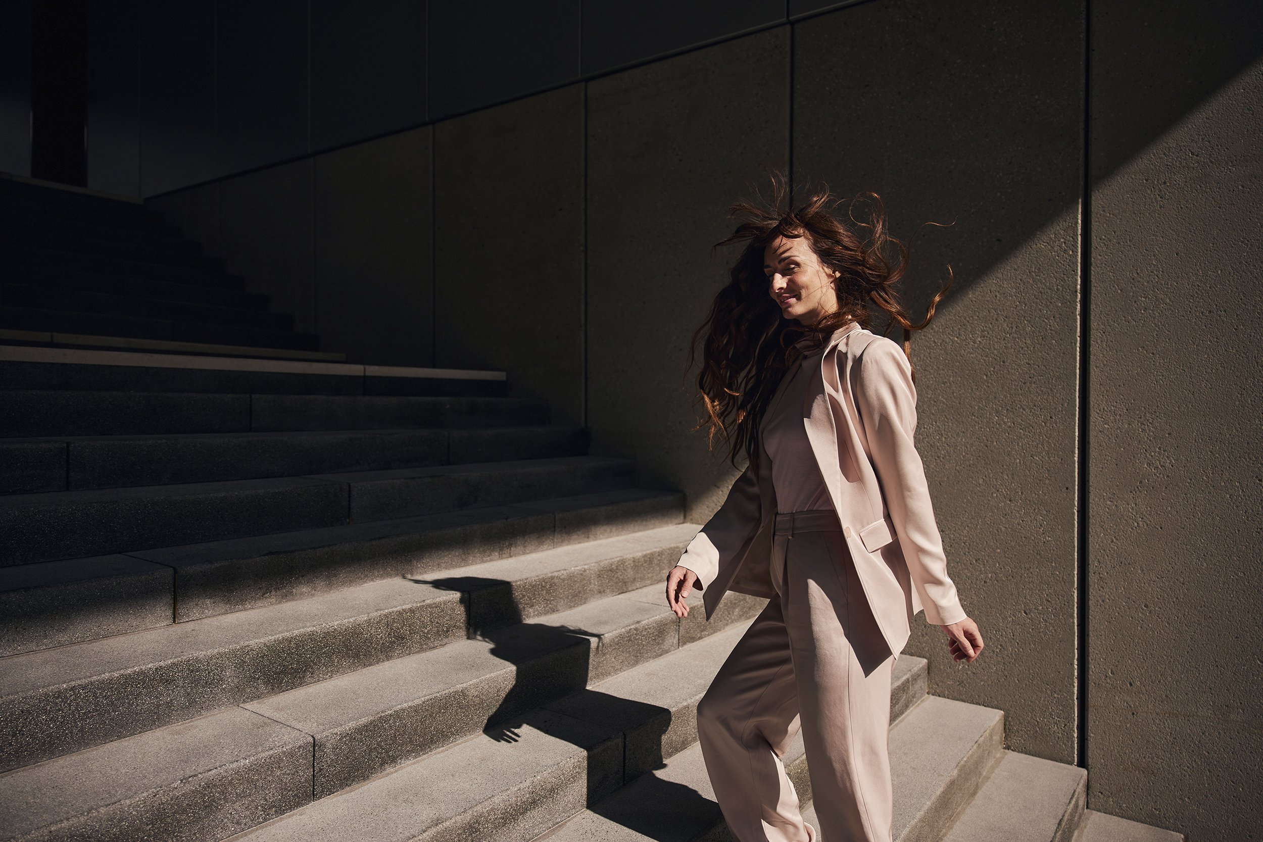 Frau in einem beige Anzug, die die Treppe hinuntergeht, bei Sonnenschein, mit Schatten auf der Wand im Hintergrund.