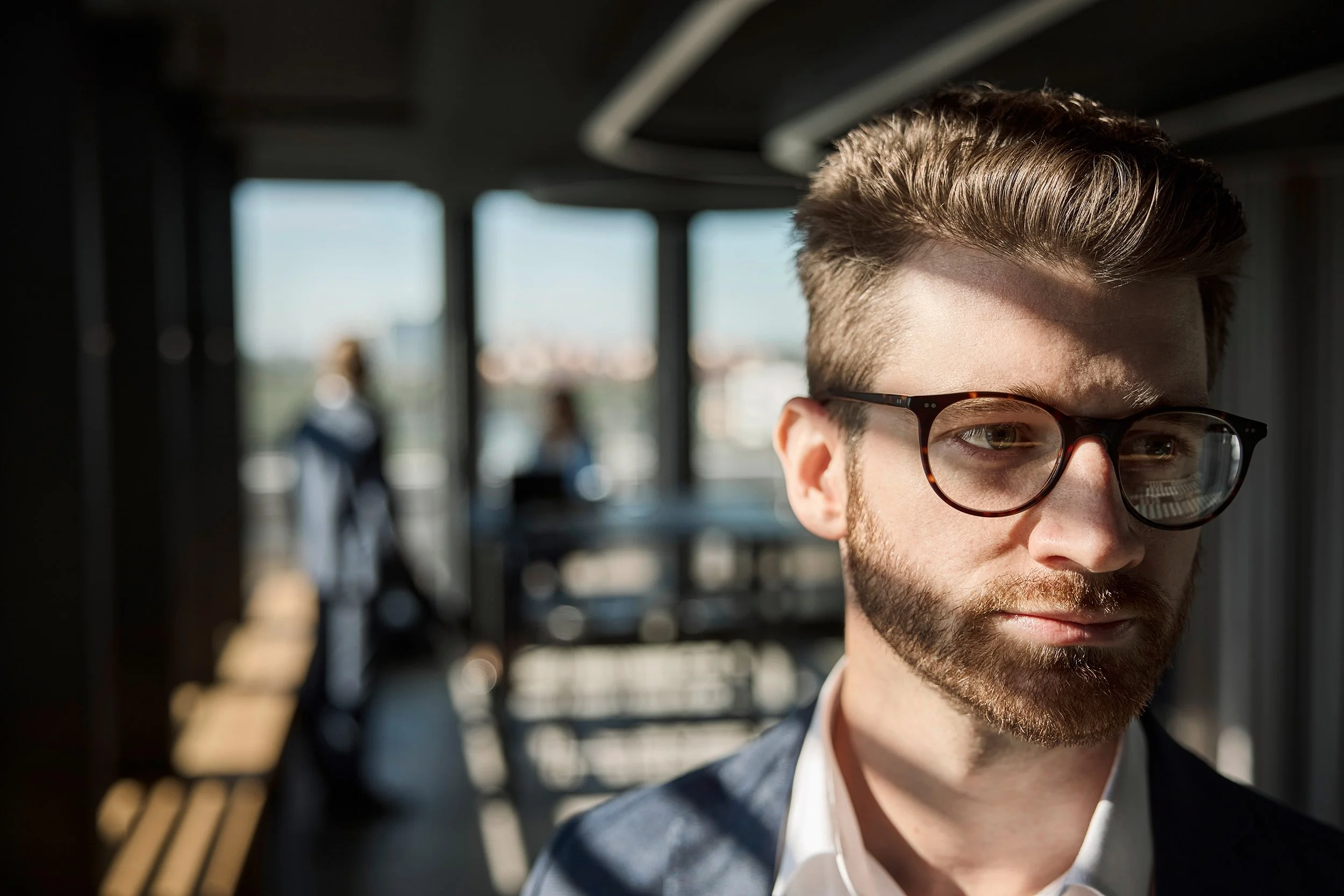 Nahaufnahme eines jungen Mannes mit Brille und Bart, der in einem helle beleuchteten Raum steht, im Hintergrund sind zwei Personen und große Fenster sichtbar.