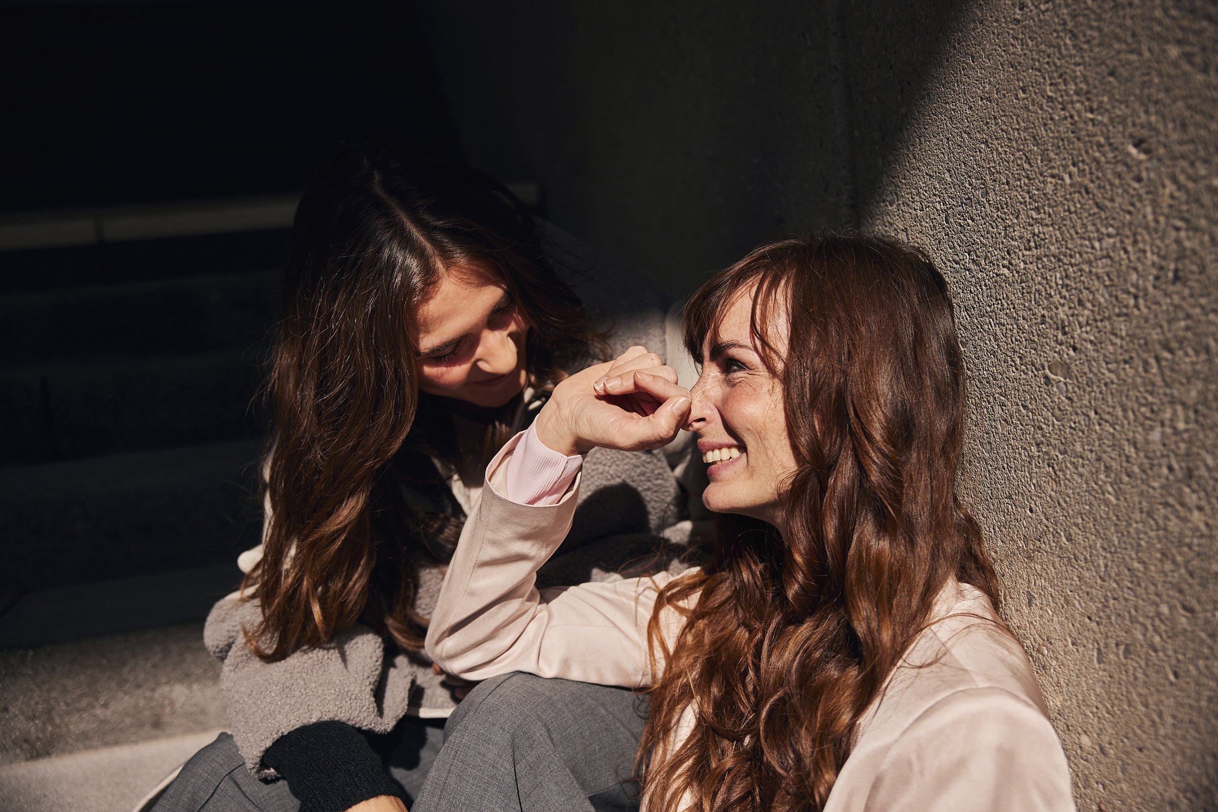 Frau mit lockigem, braunem Haar trägt eine blaue Jacke und steht in einem urbanen Umfeld vor einer modernen Architektur.