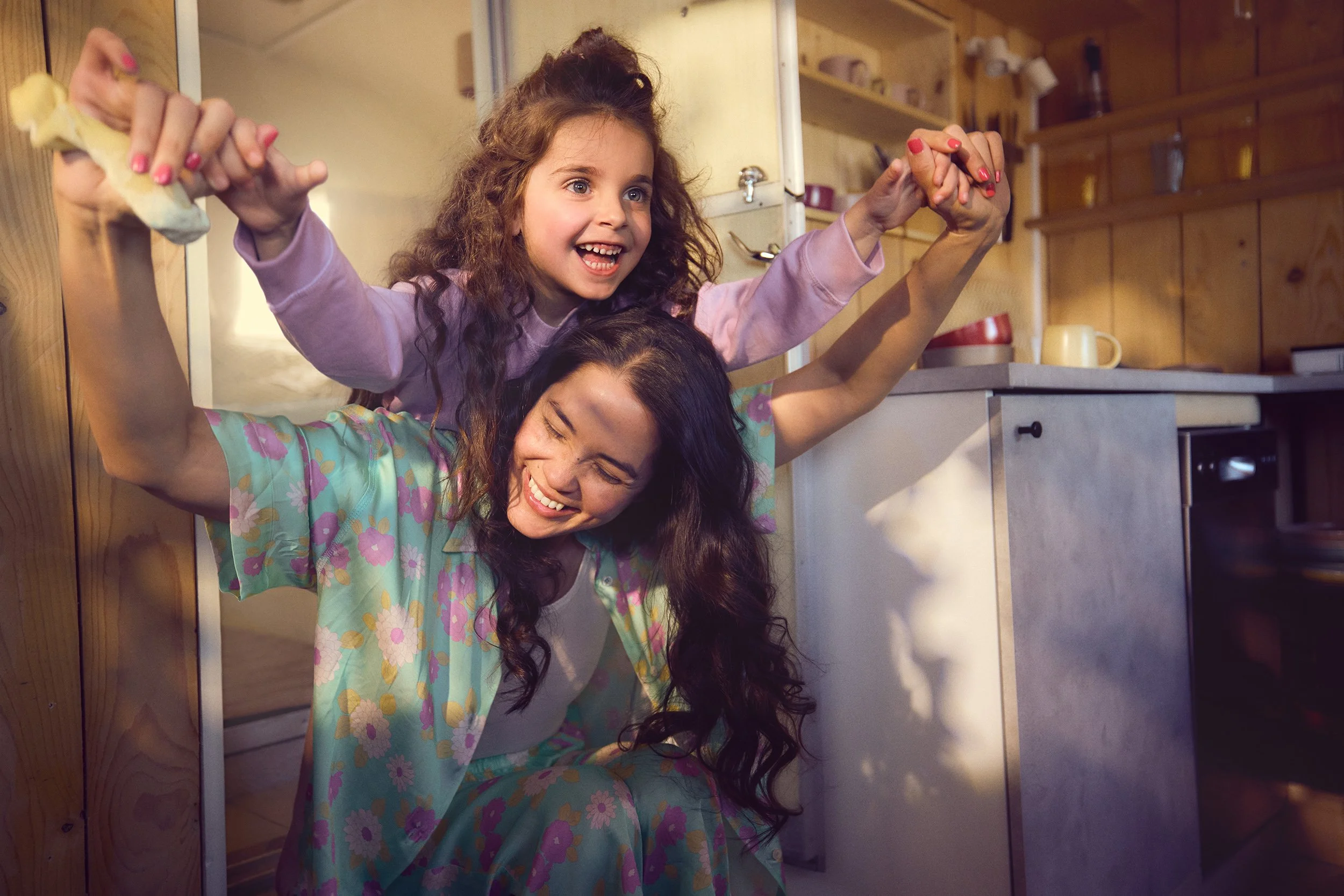 Zwei Frauen spielen fröhlich im Küchenraum, eine junge Frau mit dunkelbraunen, lockigen Haaren sitzt auf dem Boden, die andere, ein Mädchen mit großen blonden, lockigen Haaren, sitzt auf den Schultern der jüngeren, beide lachen und halten sich an den