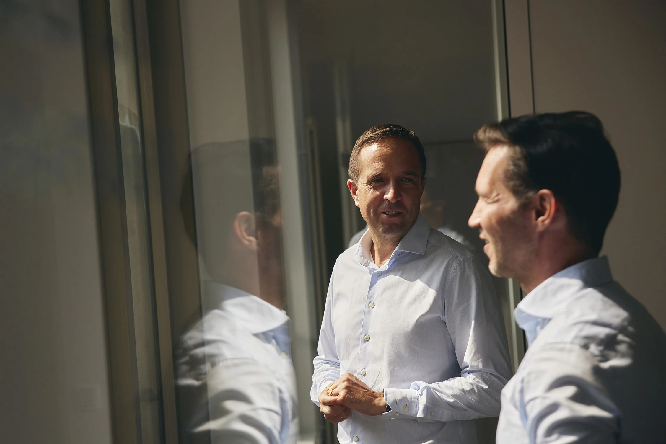Zwei Männer in weißen Hemden sprechen sich auf einem Balkon in der Sonne zu, während sie sich an einer Scheibe spiegeln.