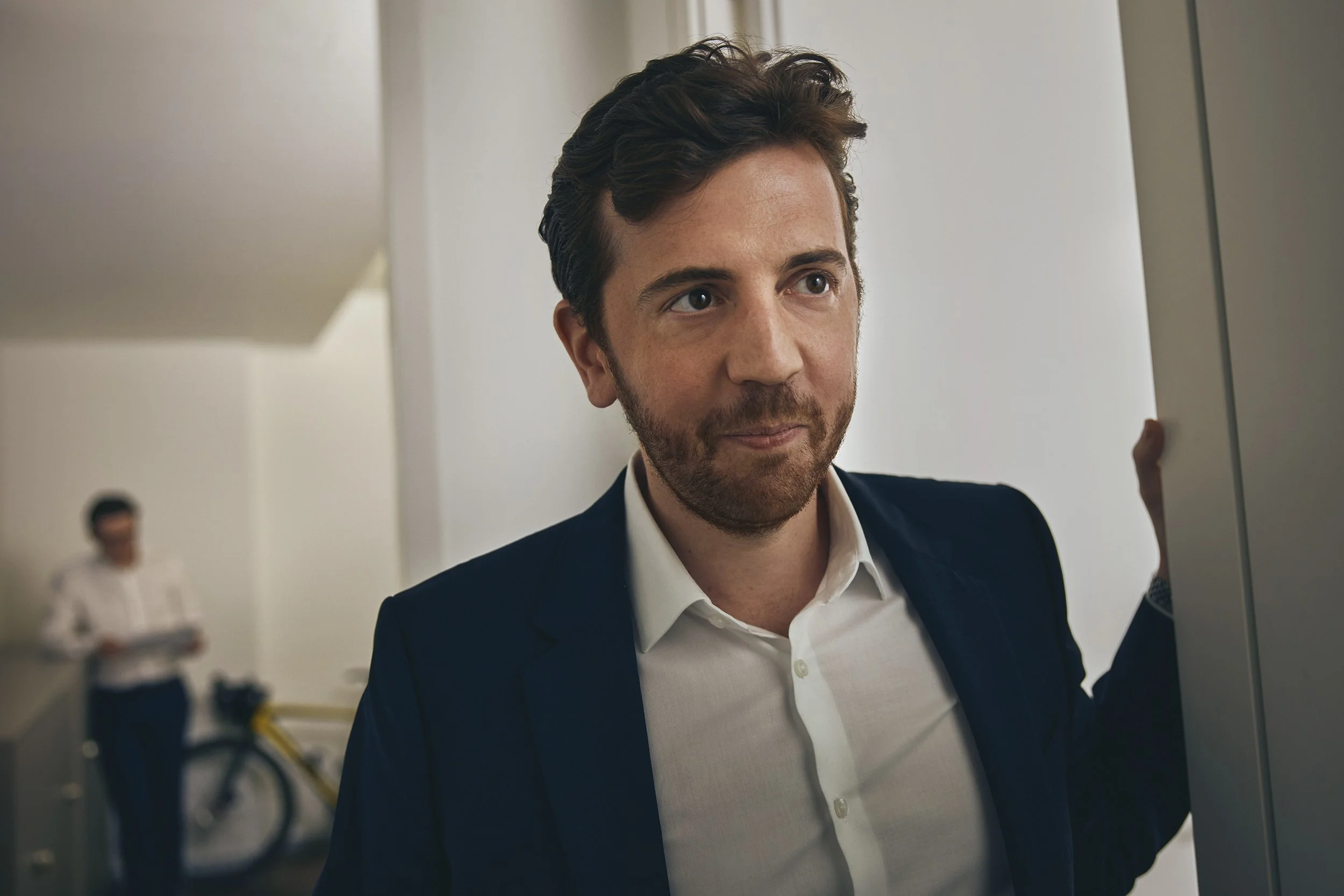 Mann in dunklem Anzug und weißen Hemd lehnt an einer Wand in einem Büro, im Hintergrund eine Frau, die ein Tablet hält und ein Fahrrad