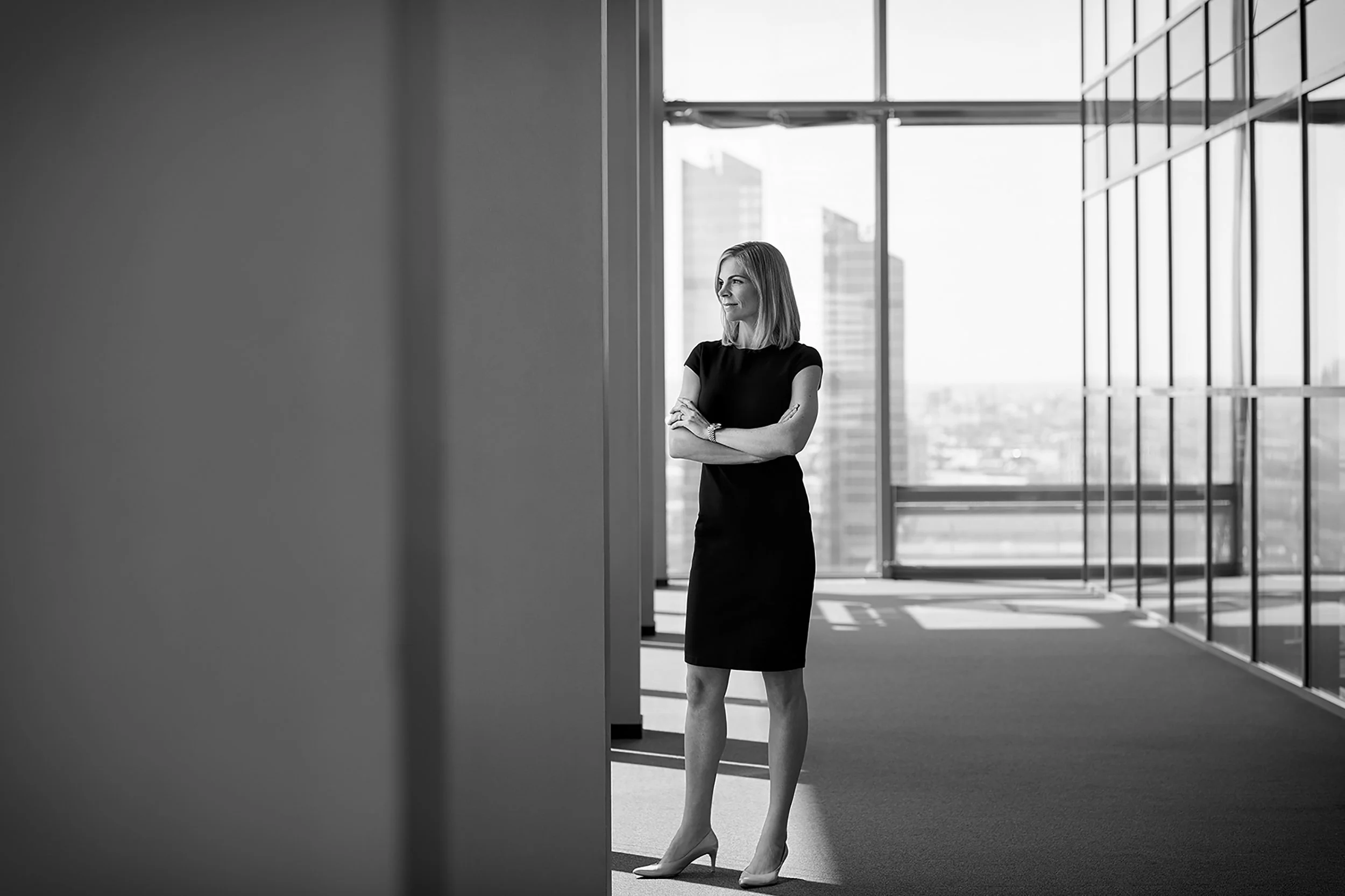 Schwangere Frau in Businesskleidung steht in einem modernen Büro mit Glaswänden und großen Fenstern, blickt nach rechts, in schwarz-weiß.