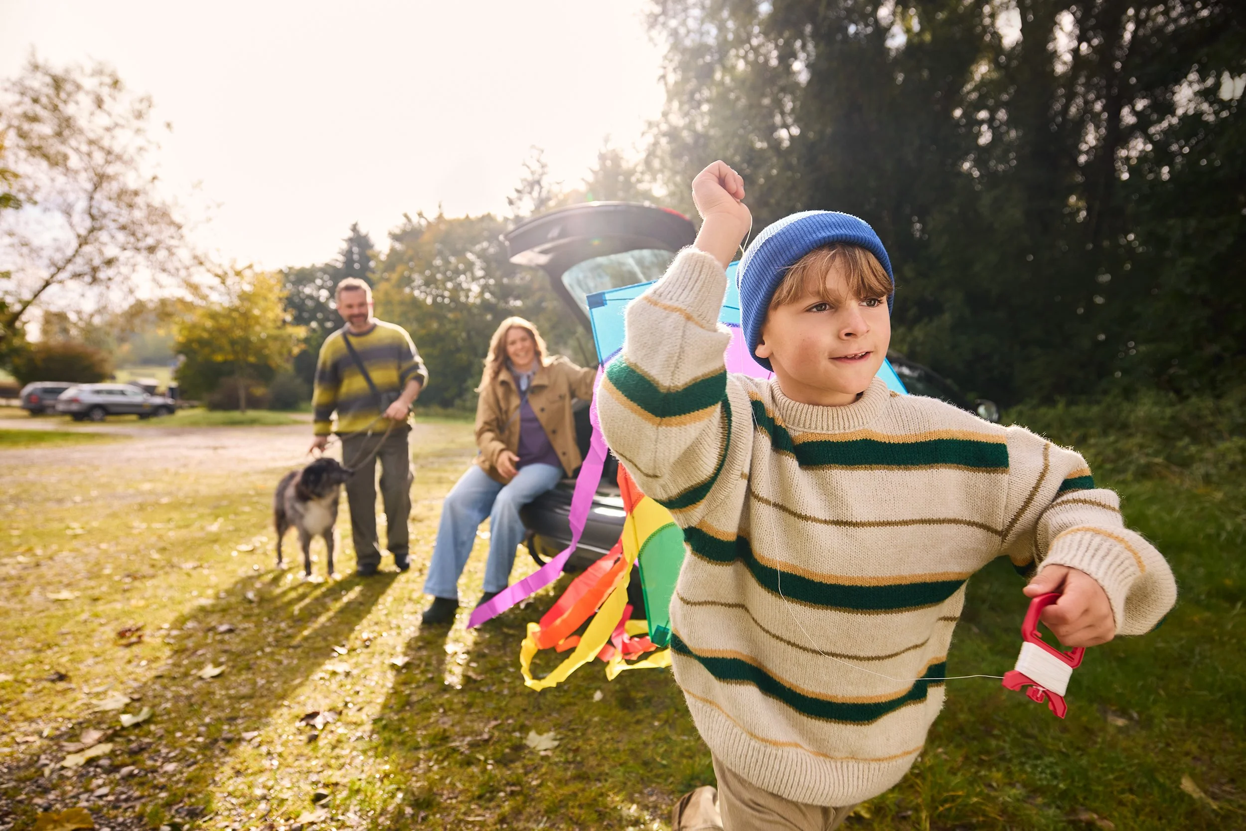 Ein Junge läuft mit einer Drachenfigur, während im Hintergrund eine Familie mit Hund im Park spazieren geht.